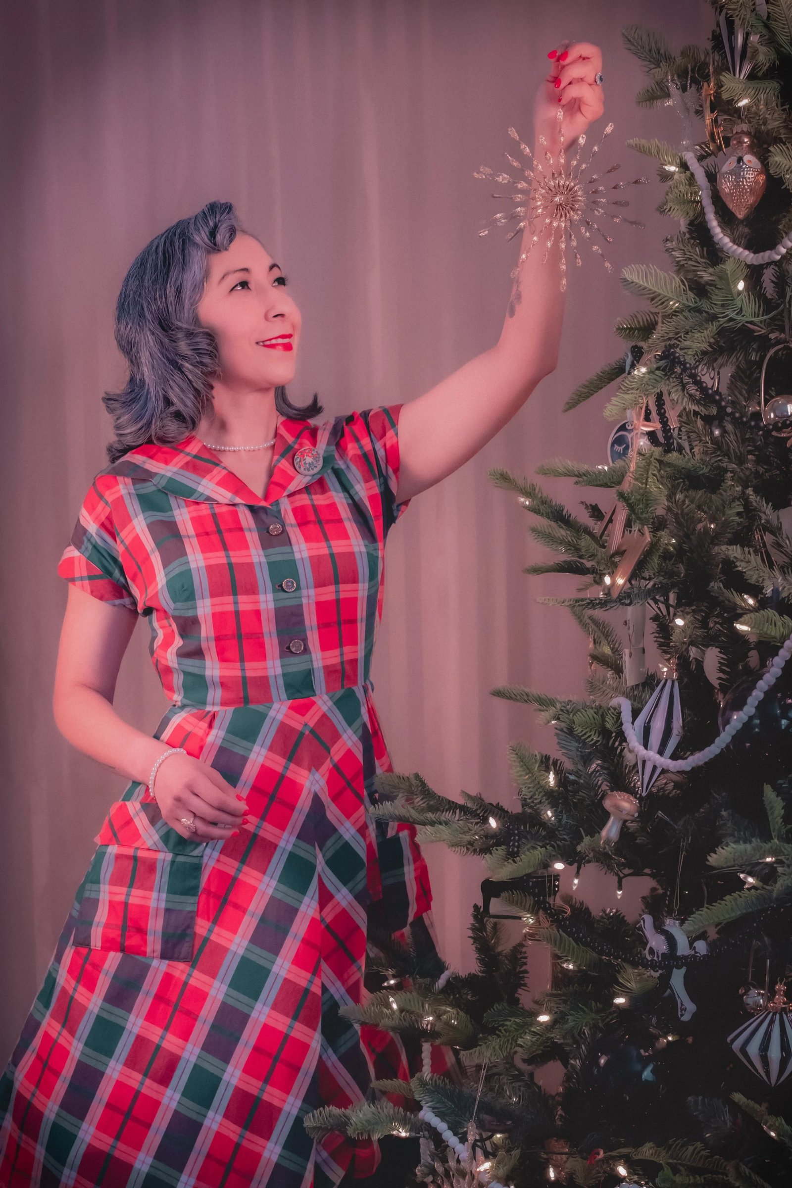 A woman in a red and green plaid dress decorates a Christmas tree.