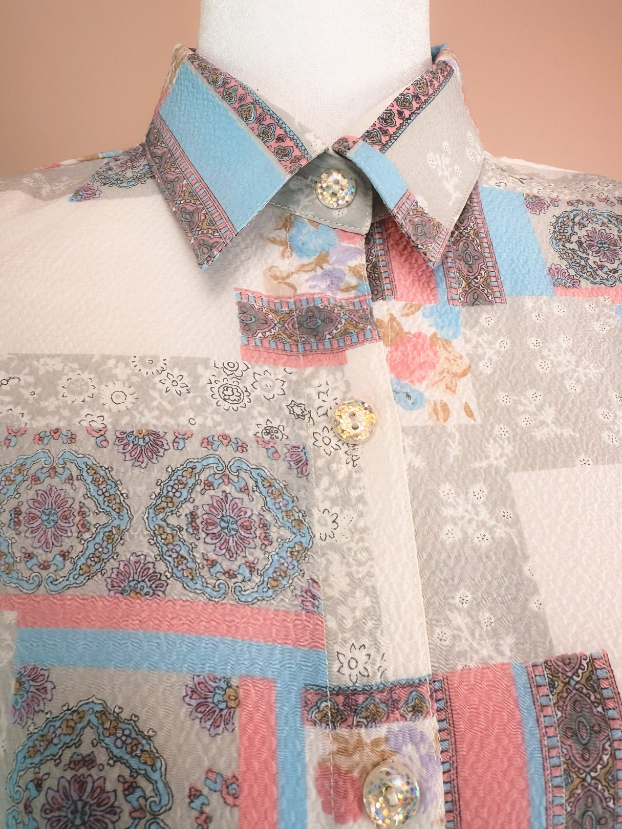 White patchwork medley blouse with intricate floral and geometric patterns, featuring a classic collar and decorative buttons, styled for women in the 1980s.