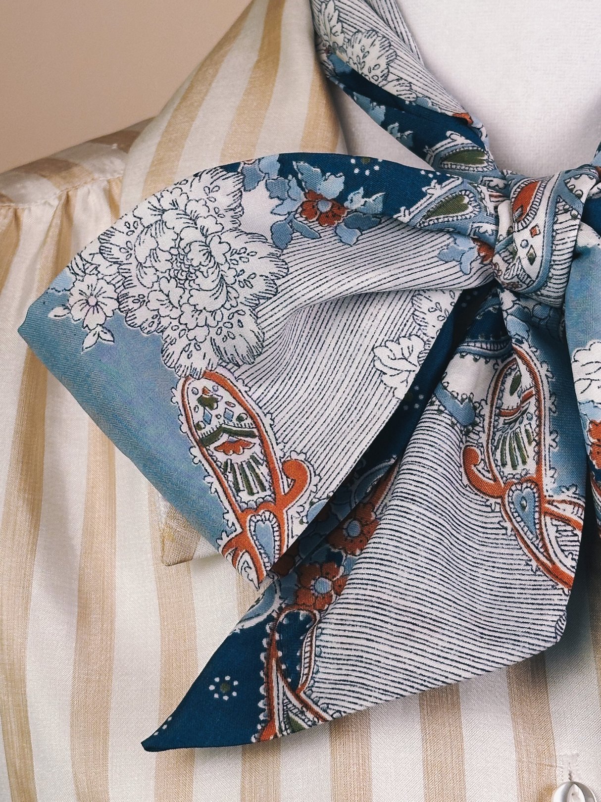 Light blue paisley scarf featuring floral and fish motifs, tied in a bow, styled with a striped blouse.