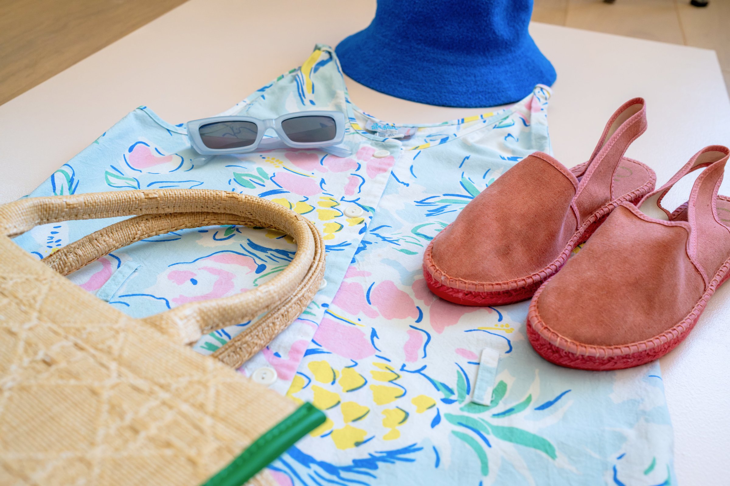 Inspired by Summer, Styled by Music gallery image: Floral-patterned blouse, blue hat, gray sunglasses, woven tote bag, and pink espadrille shoes displayed on a neutral surface.