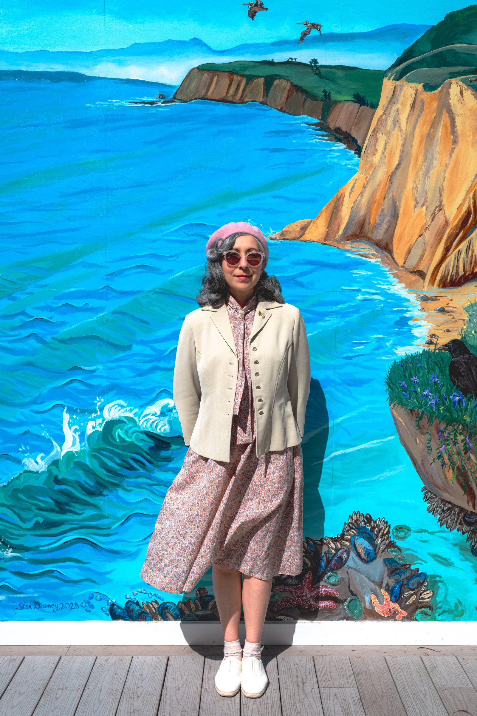 The Road to Bodega Bay gallery image: A woman in a light jacket and pink beret stands in front of a vibrant mural depicting a coastal scene with waves and cliffs.