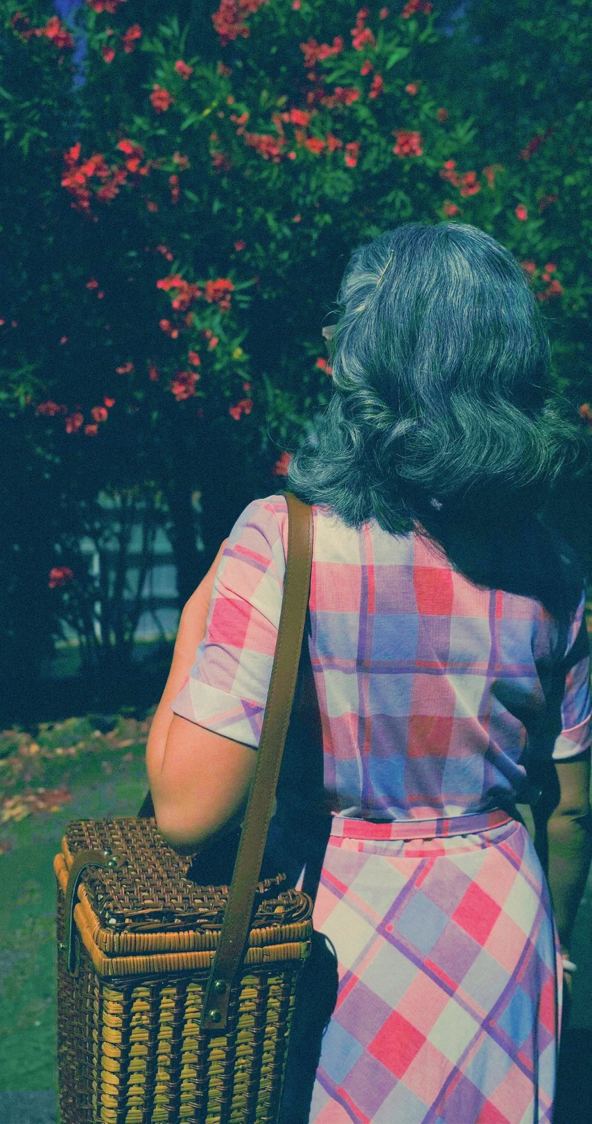 A woman with wavy gray hair in a plaid dress carries a woven basket while standing in front of flowering bushes.