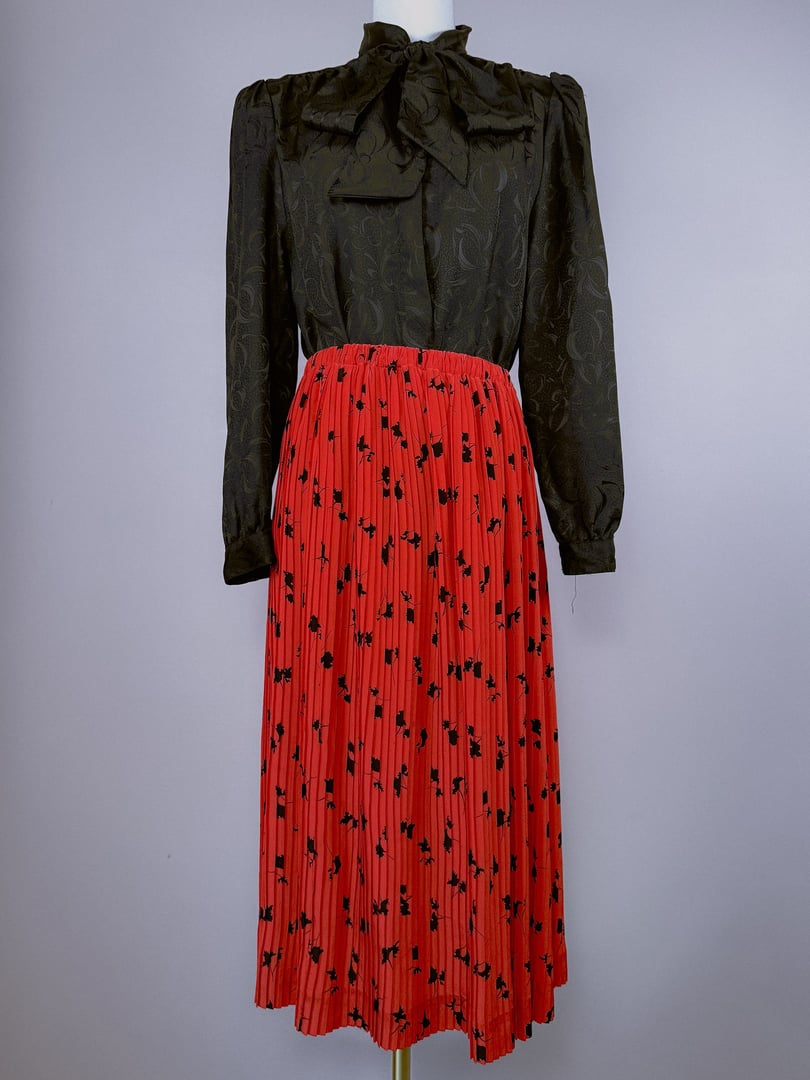 A red pleated skirt featuring a floral pattern, styled with a black long-sleeve blouse and a bow tie collar, reflecting a 1970s women's fashion aesthetic.
