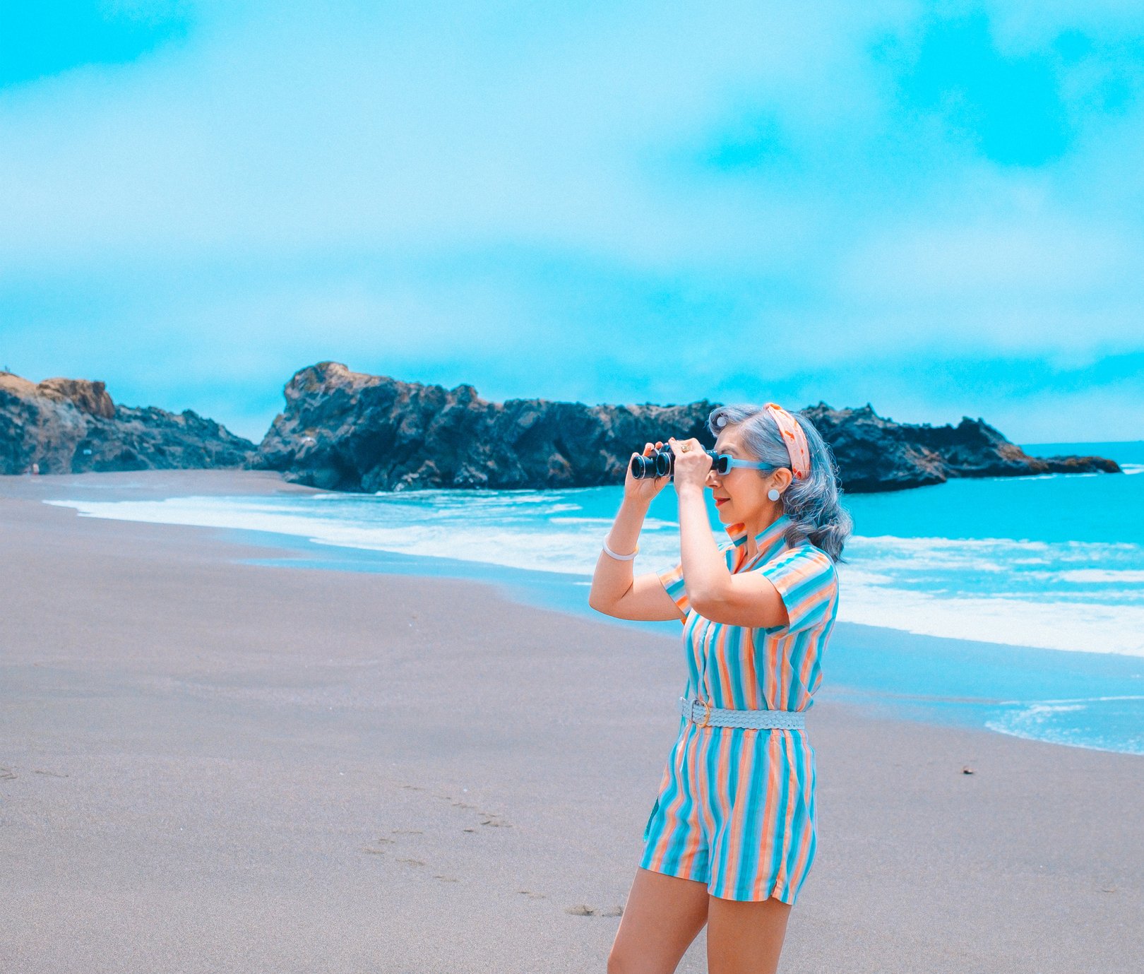 A woman in a striped romper and headband stands on a beach, holding binoculars and looking out at the ocean and rocky coastline.