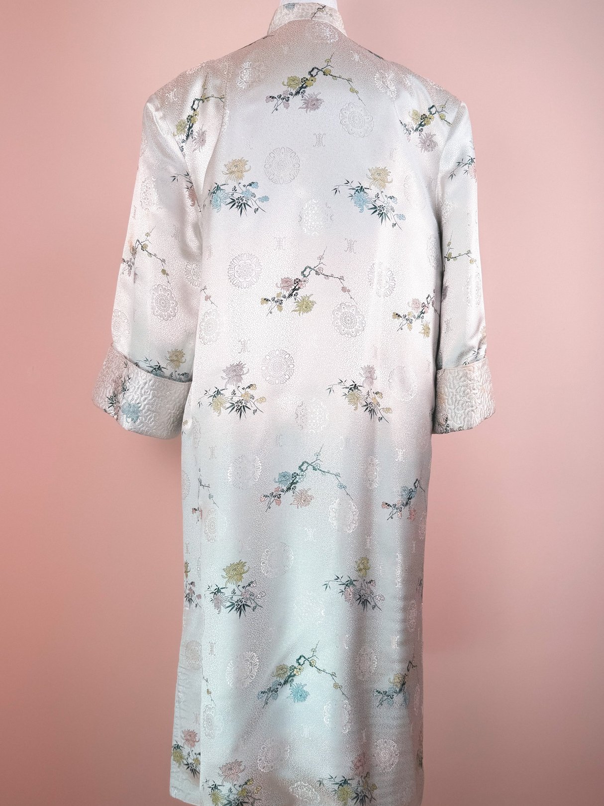Silver silk cheongsam coat for women, featuring floral patterns and 1970s design elements, with long sleeves and a knee-length cut.