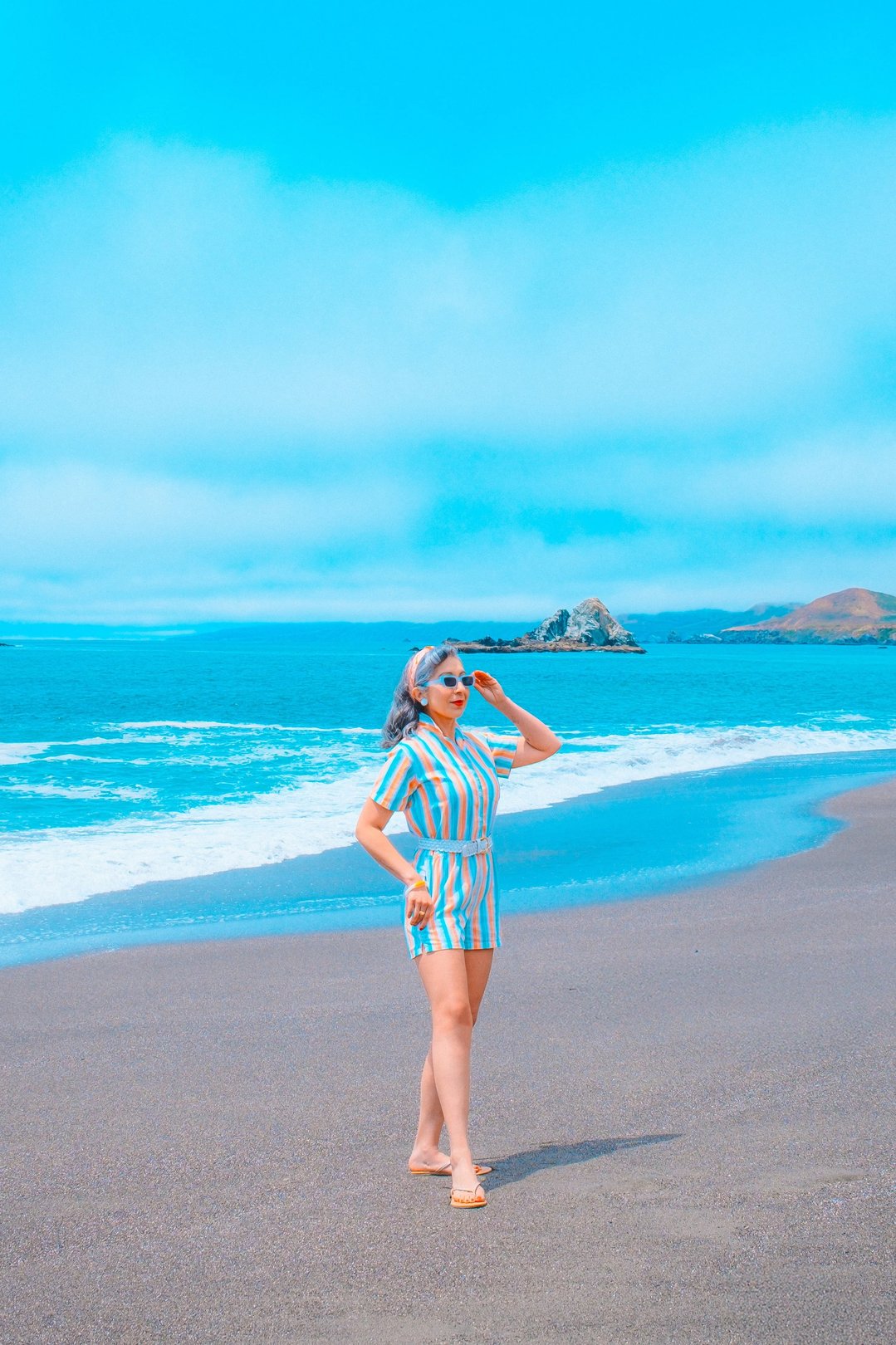 A woman in a striped beach outfit poses on a sandy shore with ocean waves and rocky formations in the background.