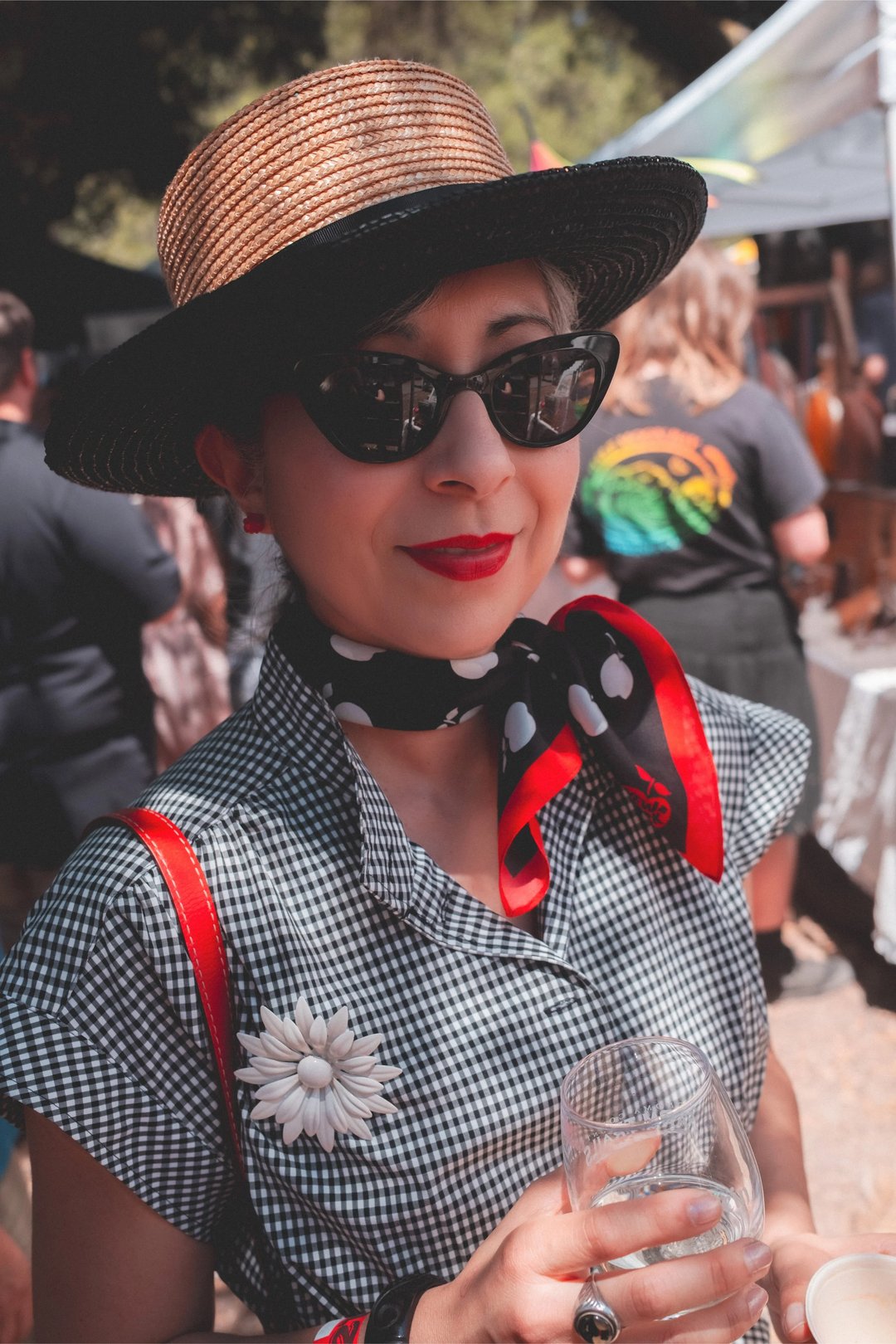 A woman wearing a wide-brimmed hat, sunglasses, and a patterned scarf poses for the camera in a crowded market.