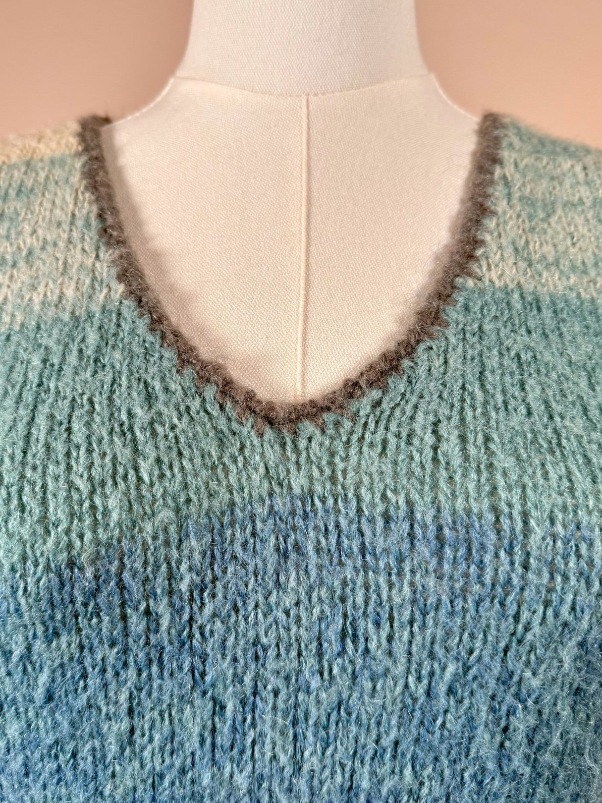 The Coastal Sky Mohair Sweater Vest features a V-neck design with a textured knit in varying shades of blue, typical of 1970s fashion.