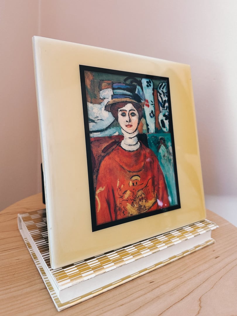 Ceramic portrait titled "La fille aux yeux verts" by Matisse, featuring a woman in a red outfit with a yellow background, displayed on a light-colored wooden table.