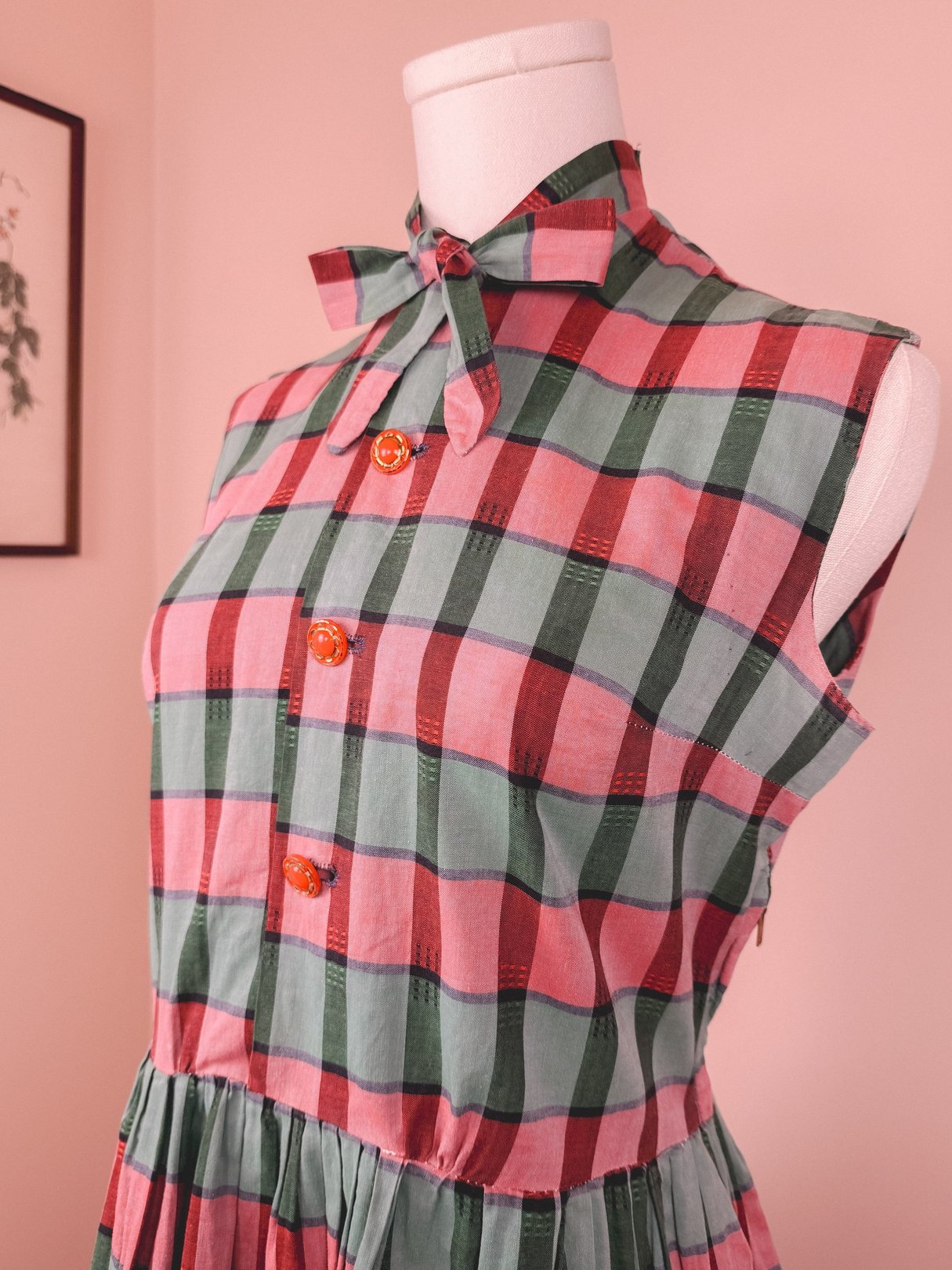 The Holiday Carol Plaid Dress features a sleeveless design with a bow tie collar, in red, green, and pink plaid, adorned with orange buttons.