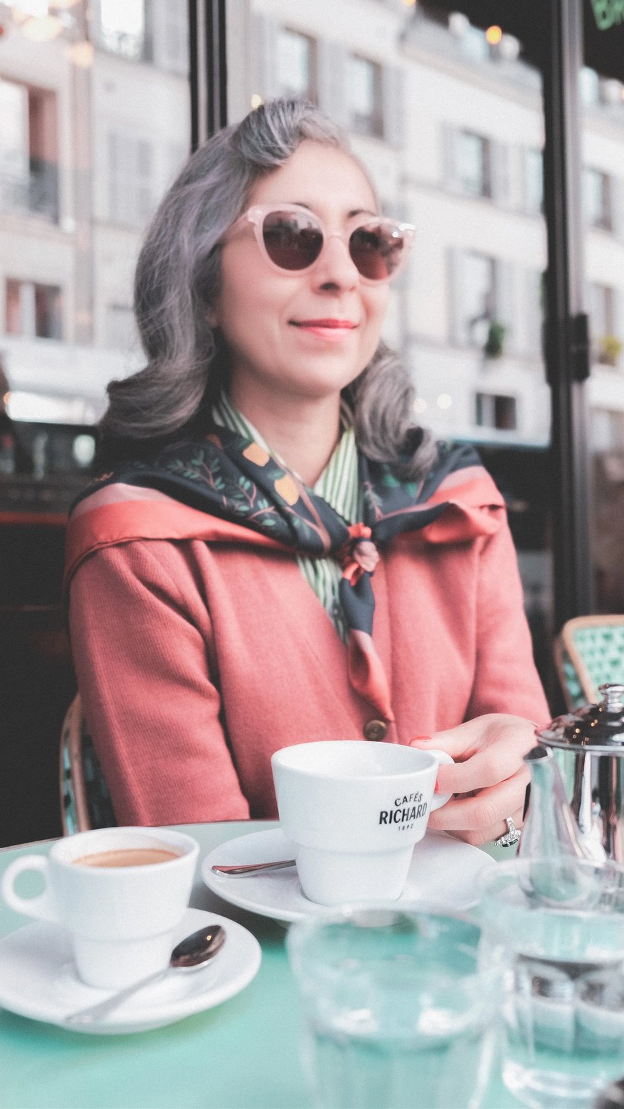 A woman with gray hair wearing sunglasses, a pink cardigan, and a patterned scarf sits at a café table with a cup of coffee and a small espresso.