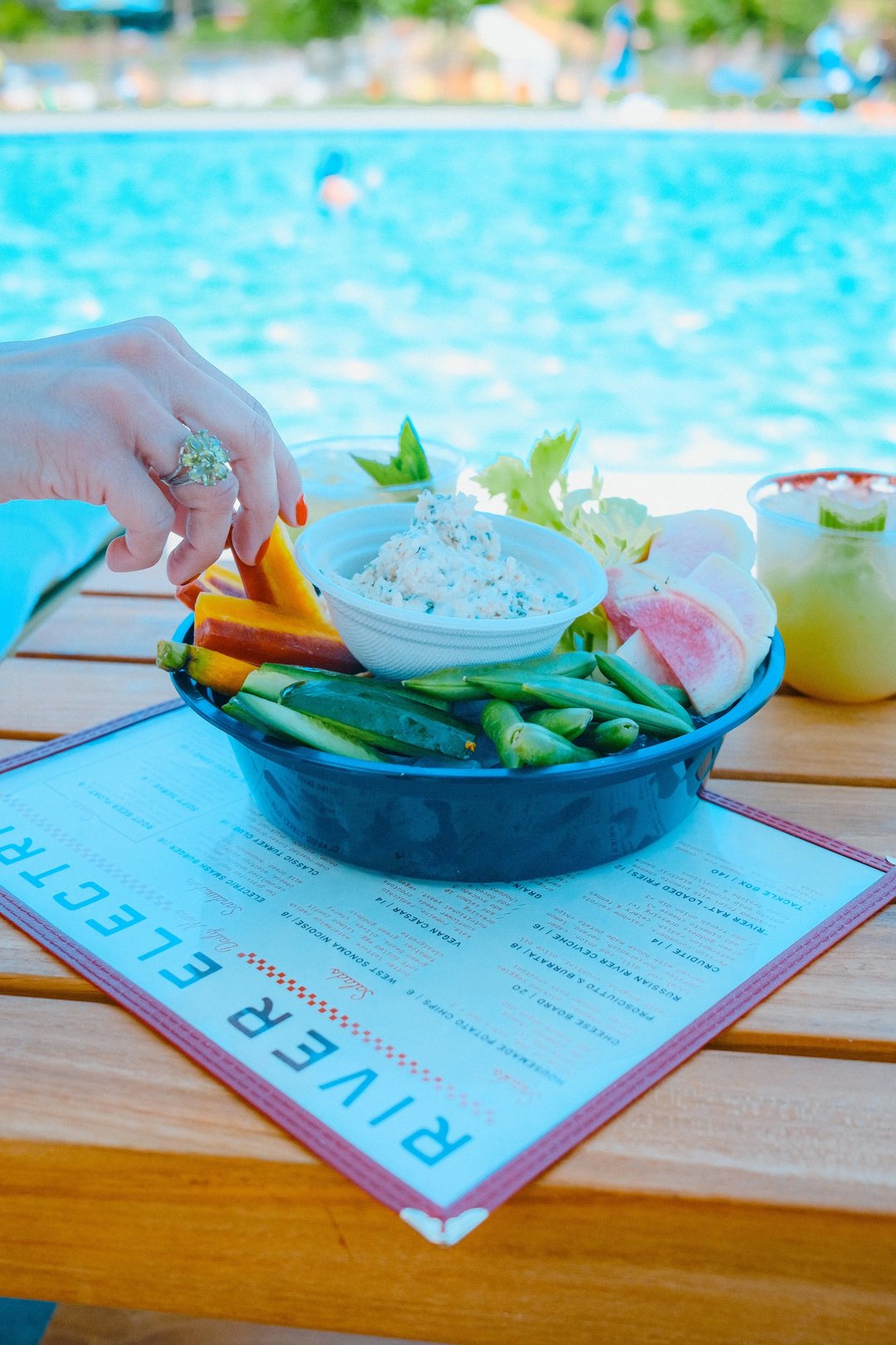 A hand reaches for a bowl of fresh vegetables and dip, accompanied by a drink, on a table by the pool.