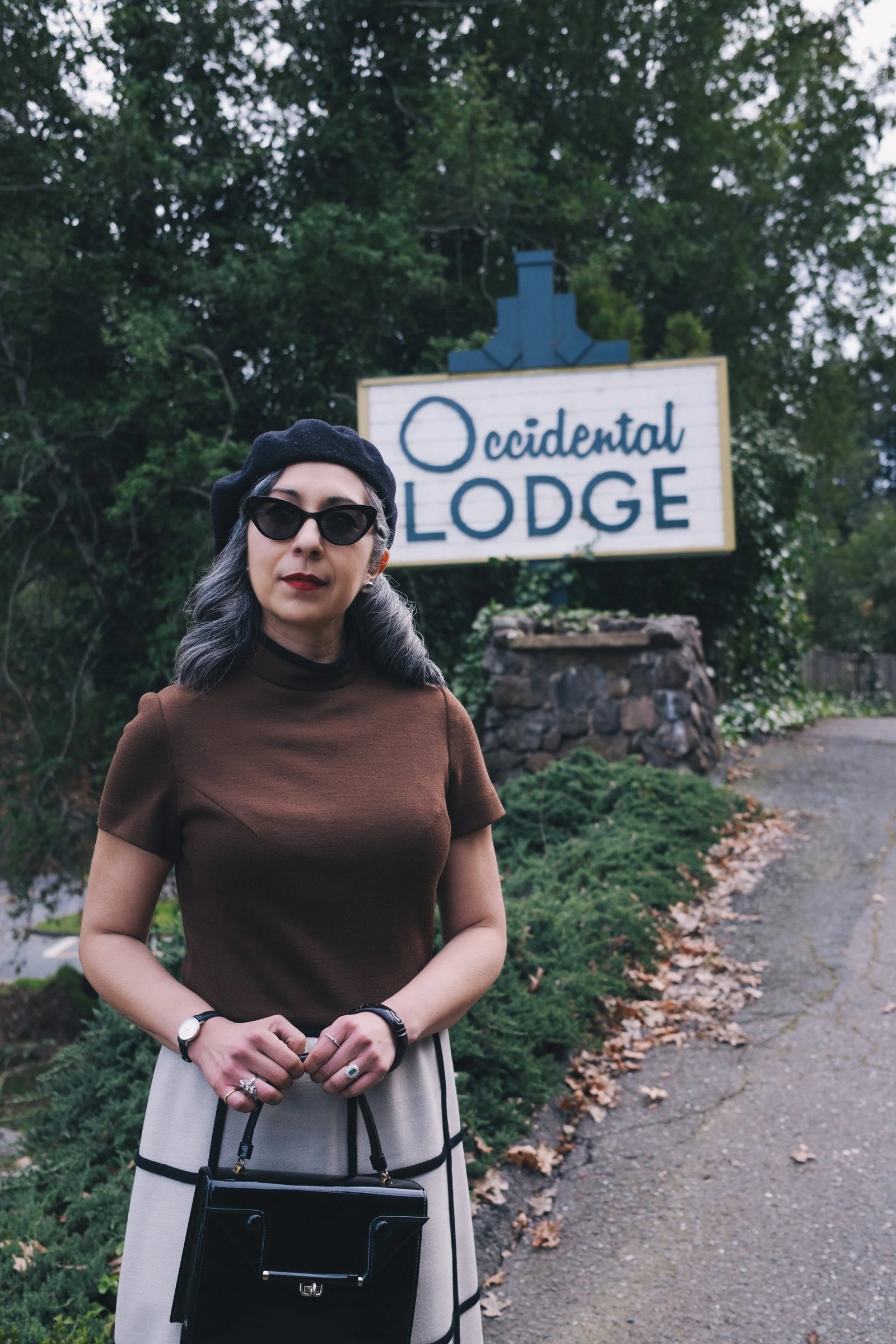 A woman with gray hair wearing a brown top, black beret, and sunglasses stands in front of the Occidental Lodge sign.