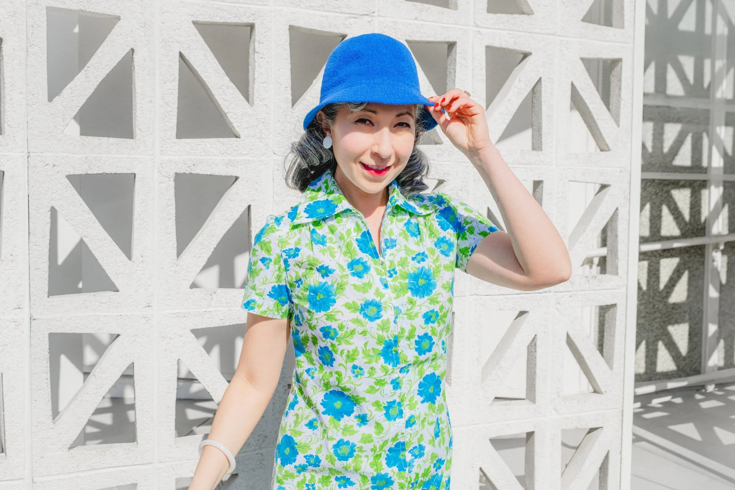 Woman wearing a blue bucket hat and a floral dress stands in front of a textured white wall, smiling at the camera.