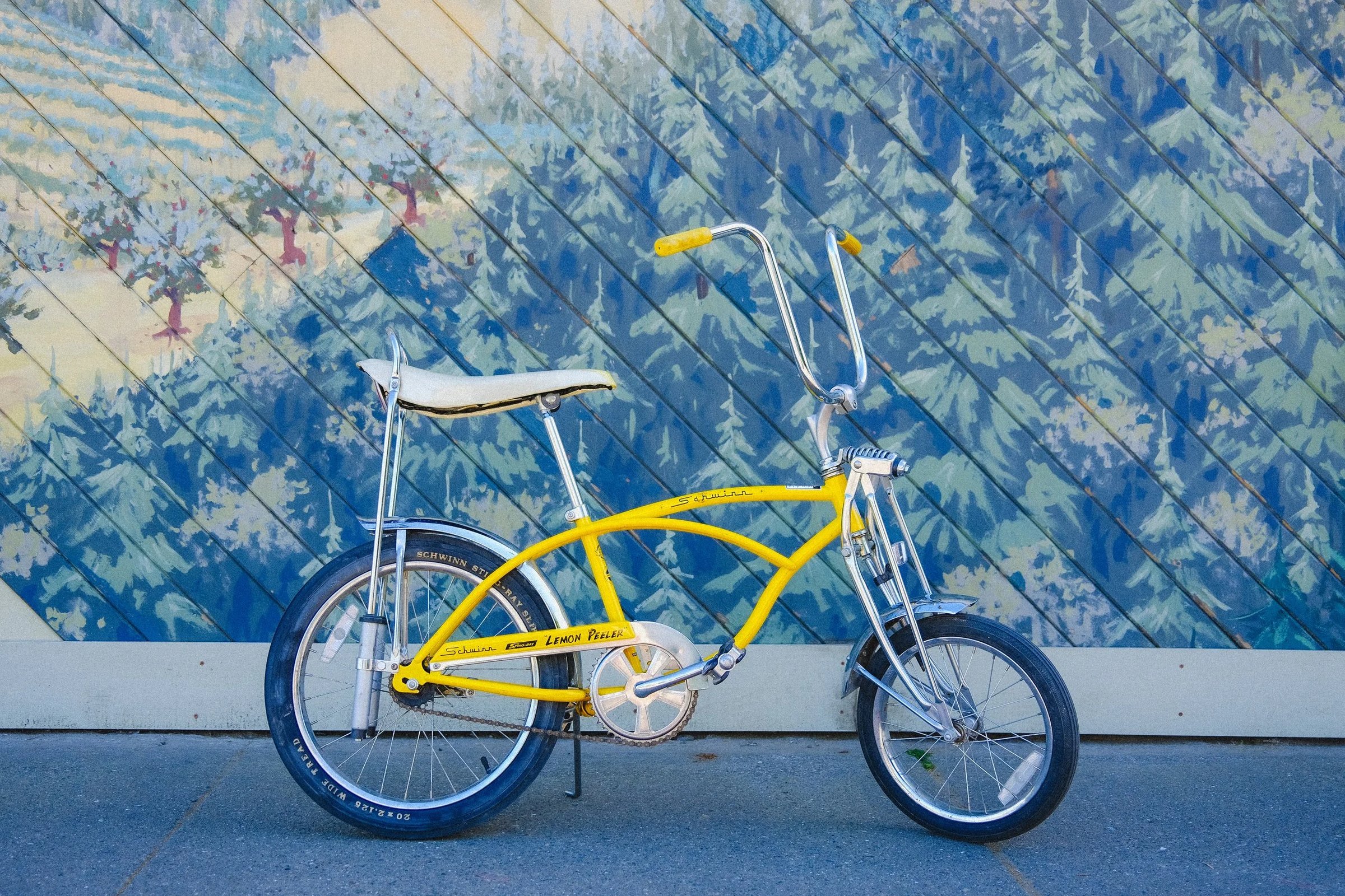 A vintage yellow bicycle with a unique frame design and a white seat, leaning against a blue painted wall.