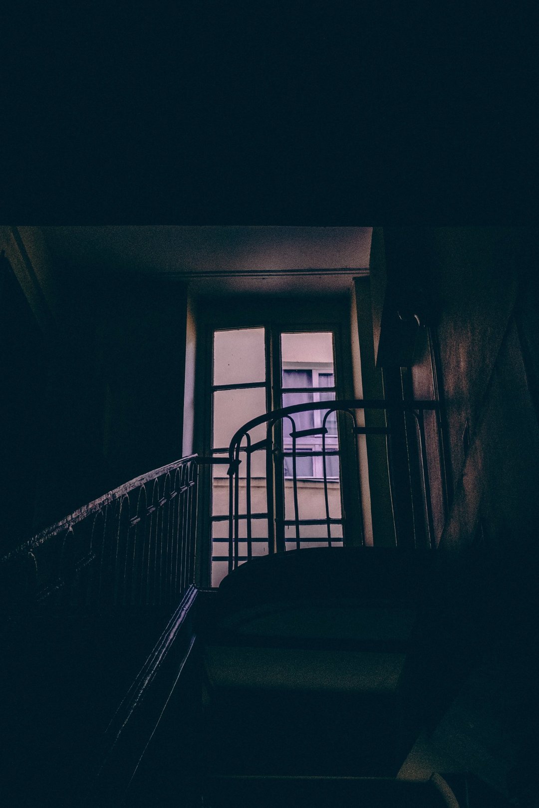 A dimly lit staircase with a metal railing leads to a window with purple curtains.
