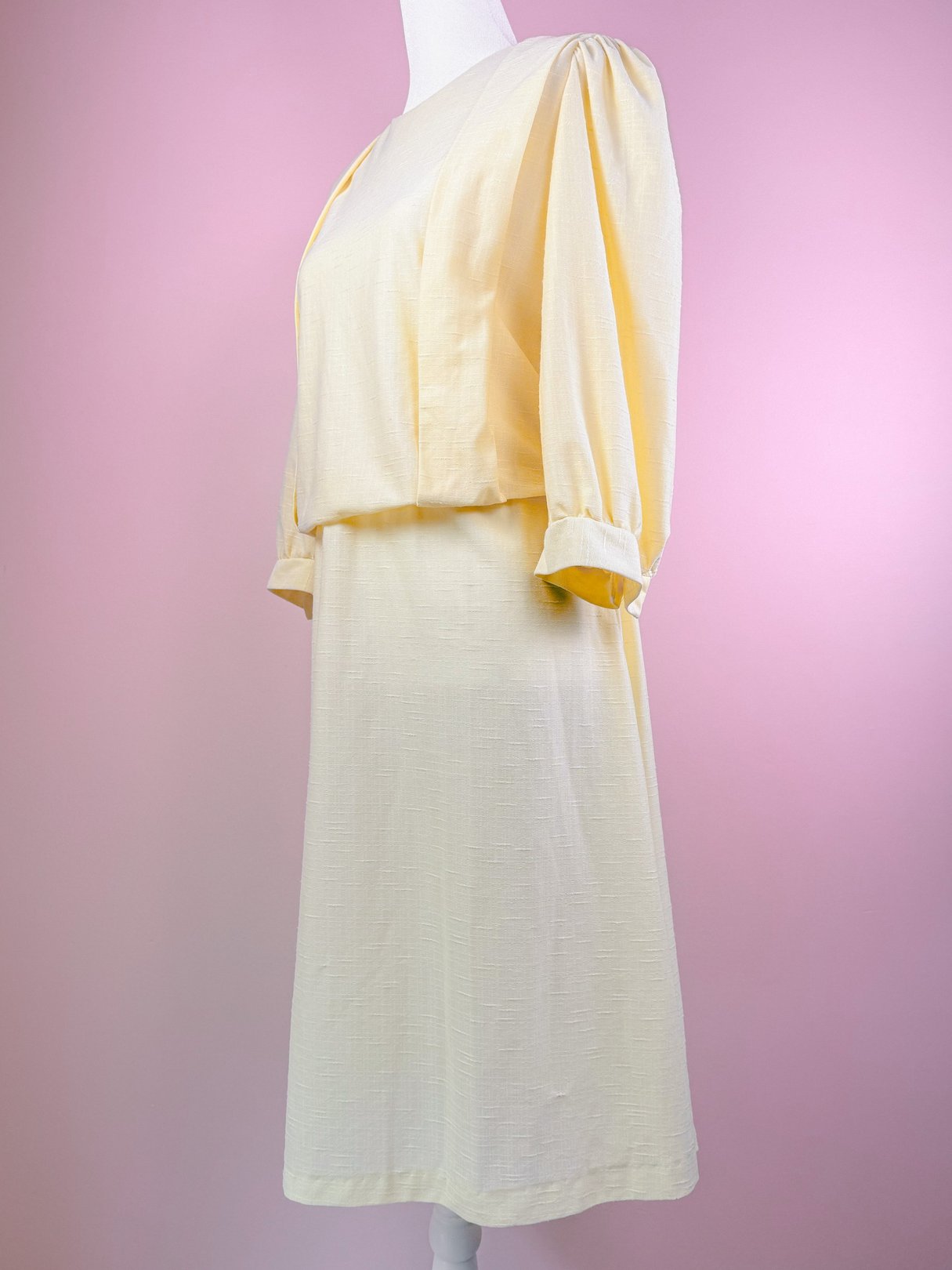 A 1980s women's lemon chiffon dress featuring a fitted bodice and loose, puffed sleeves, displayed on a dress form against a pink background.