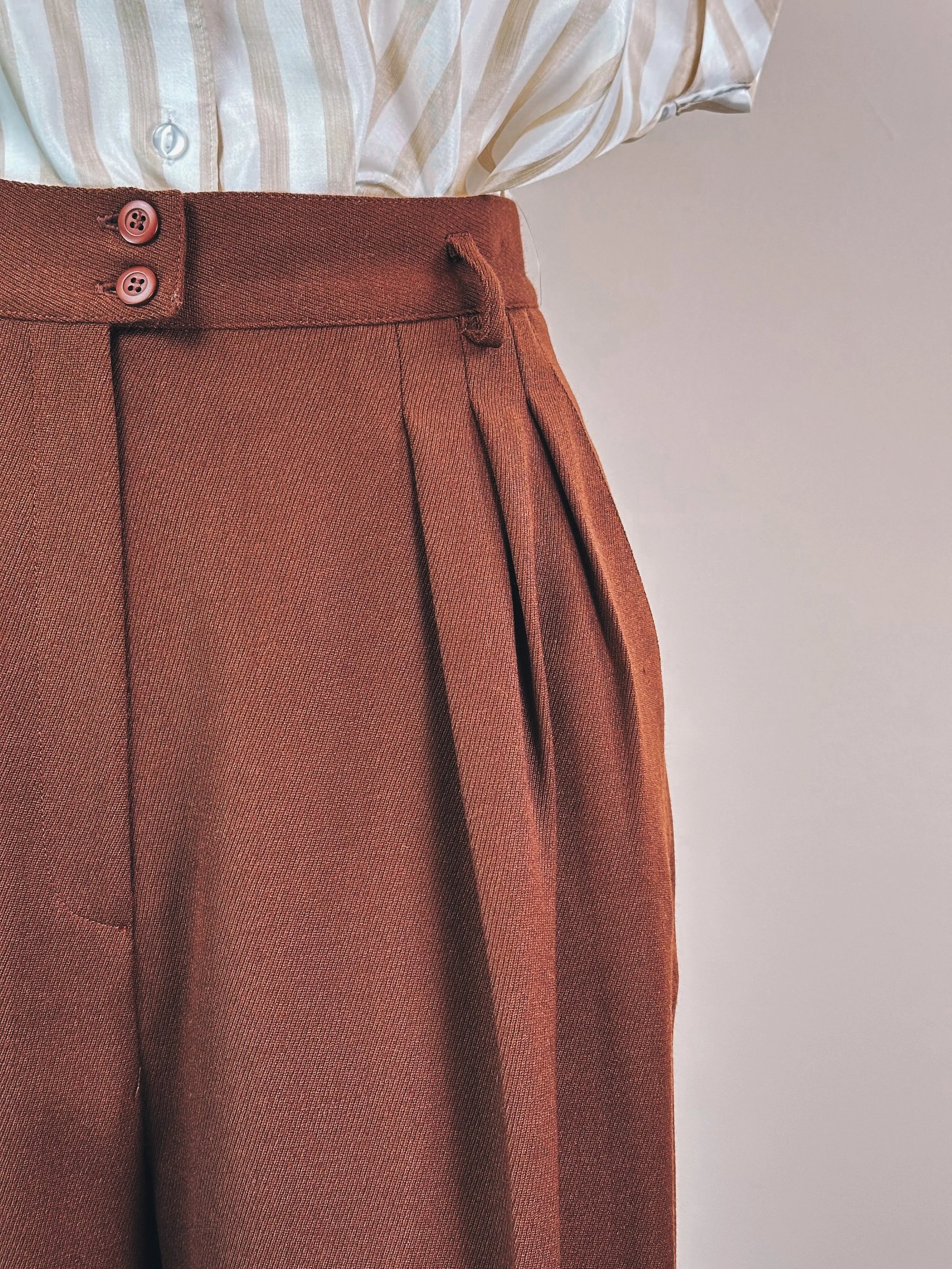 Brown pleated trousers with two buttons paired with a light striped shirt.