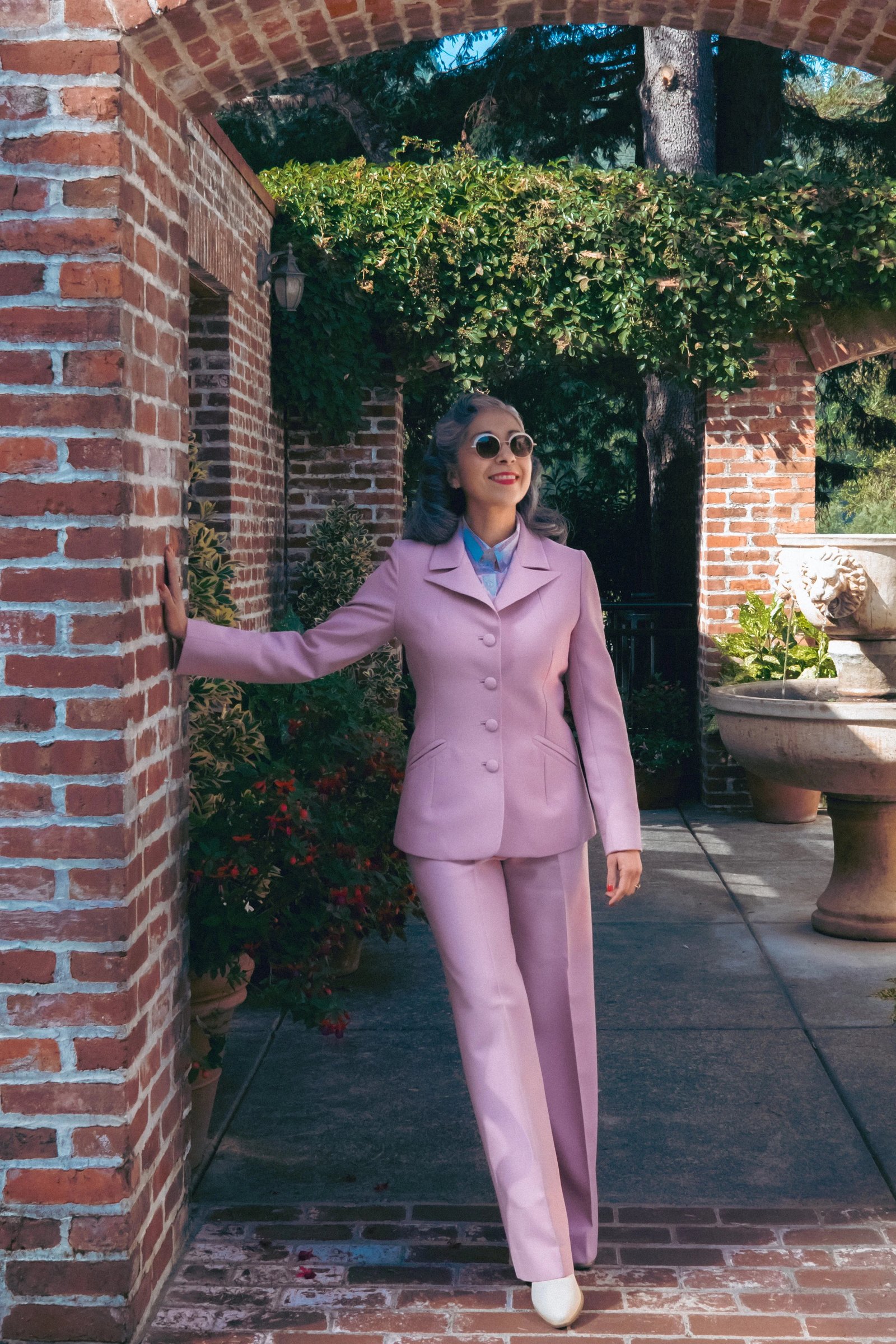 A woman wears a pink suit and sunglasses while standing outdoors near brick walls and greenery.