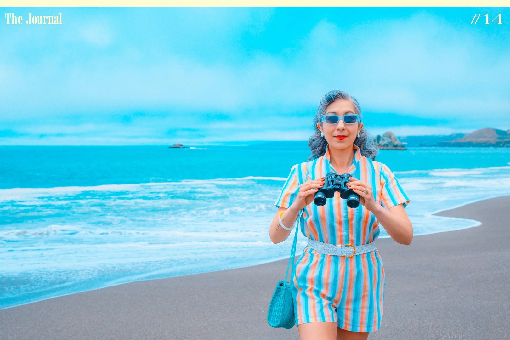 A woman in a striped romper and sunglasses holds binoculars while standing on a beach with ocean waves in the background.