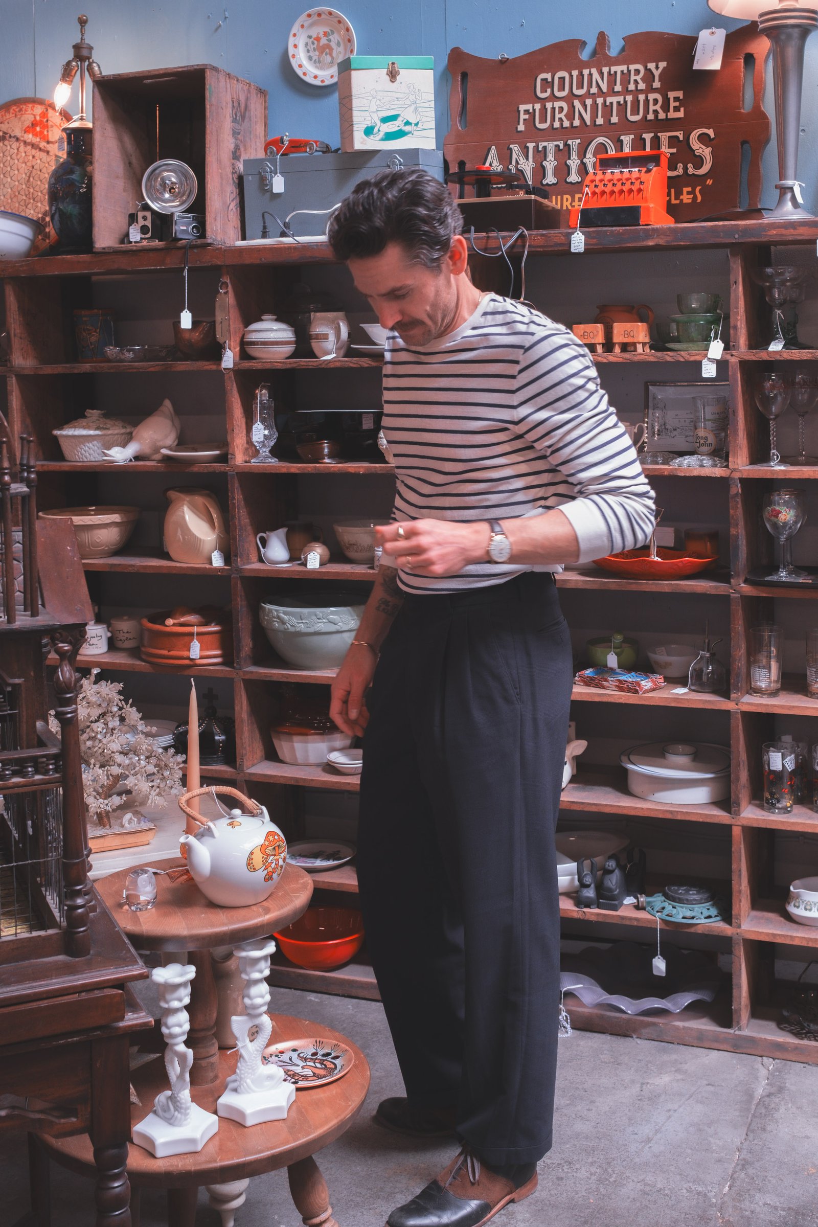 Sheet Music Stories gallery image: A person in a striped shirt and black pants stands beside a rustic wooden shelf filled with various vintage pottery and decorative items.