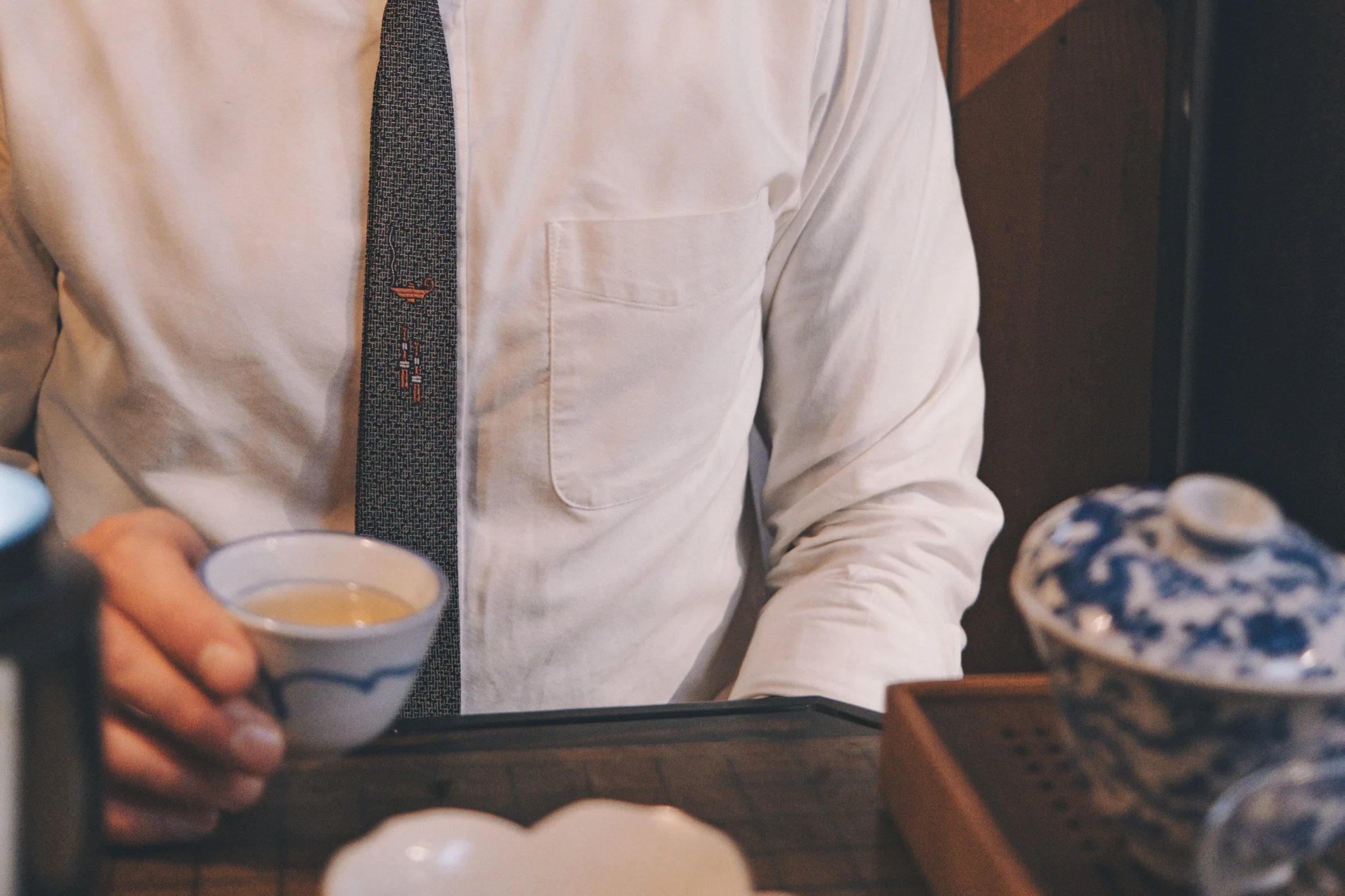 A person wearing a white shirt and patterned tie holds a small blue and white teacup.