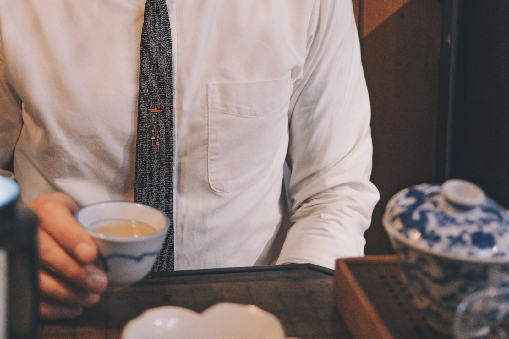 A person wearing a white shirt and patterned tie holds a small blue and white teacup.