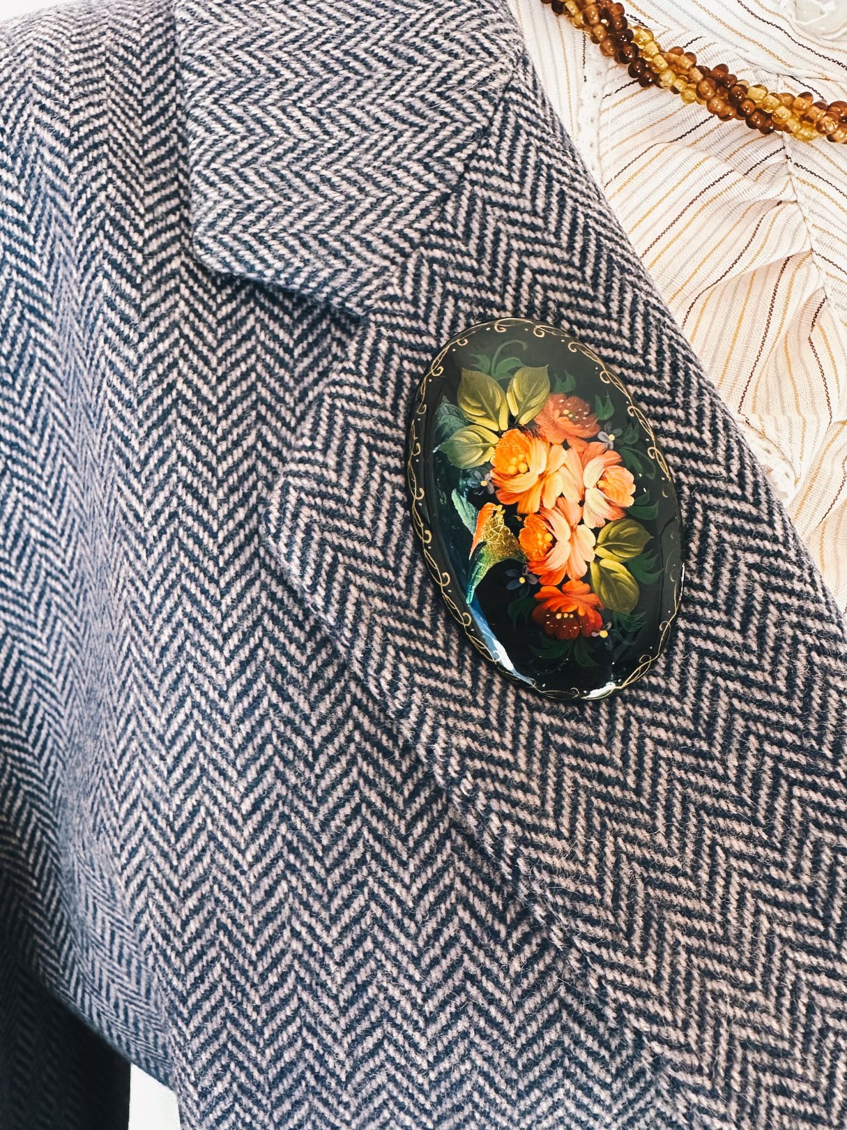 A black Fluttering Bird brooch featuring a colorful floral design, worn on a herringbone-patterned blazer.