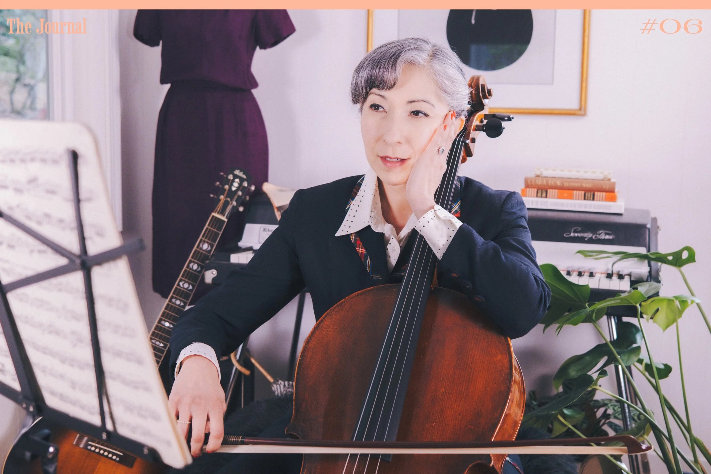 A woman with gray hair sits with a cello, wearing a blazer and a white collared shirt, in a well-decorated room.