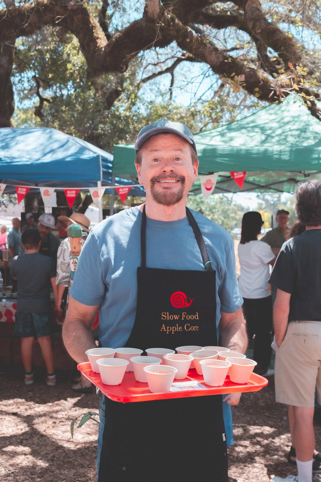A smiling man wearing an apron holds a tray of small cups at an outdoor food event with tents in the background.
