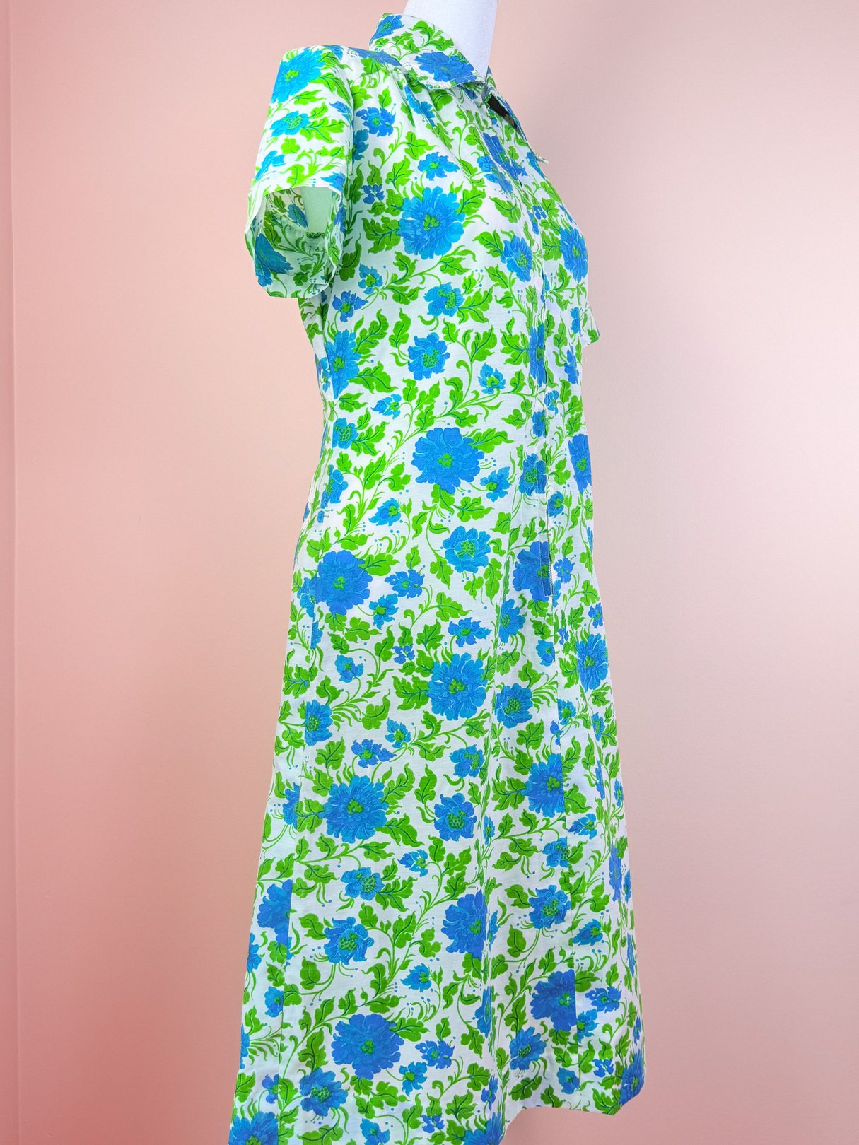 1960s women's blue flower print dress featuring short sleeves and a knee-length silhouette against a pink background.