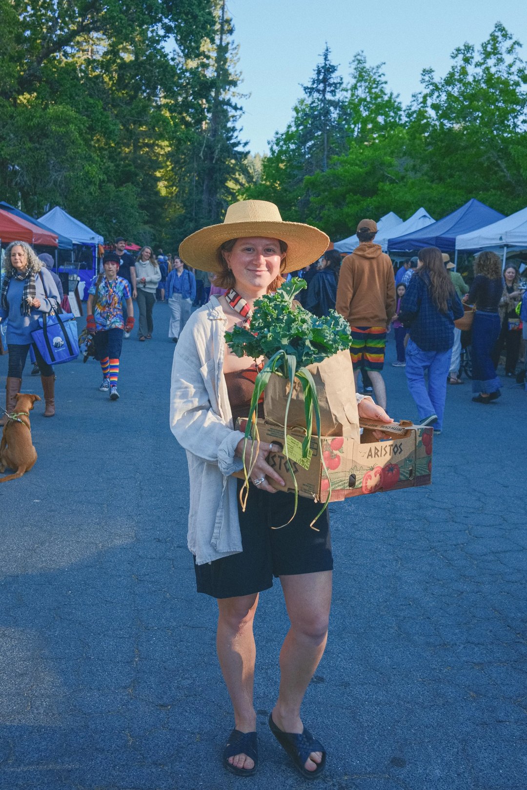 A woman wearing a wide-brimmed hat holds a box of fresh kale at a farmer's market while people browse in the background.