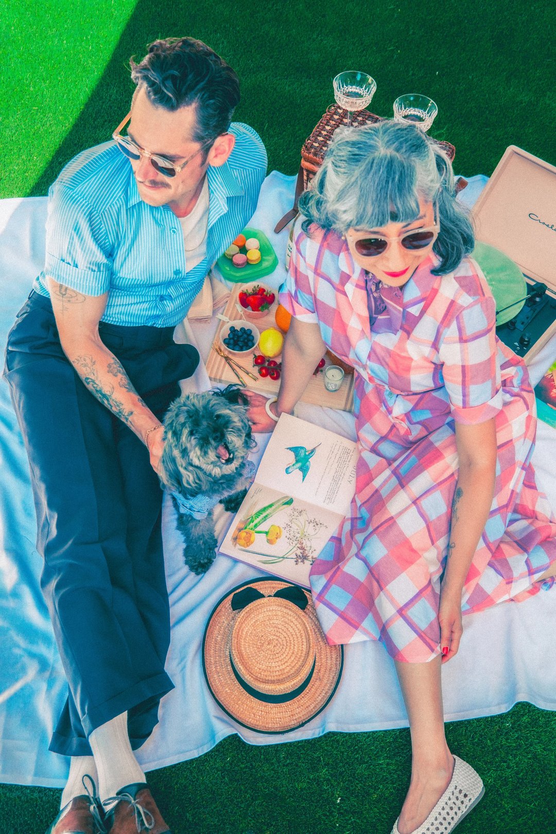 A couple enjoys a picnic on a sunny day, surrounded by colorful snacks and a small dog, dressed in vintage-inspired outfits.