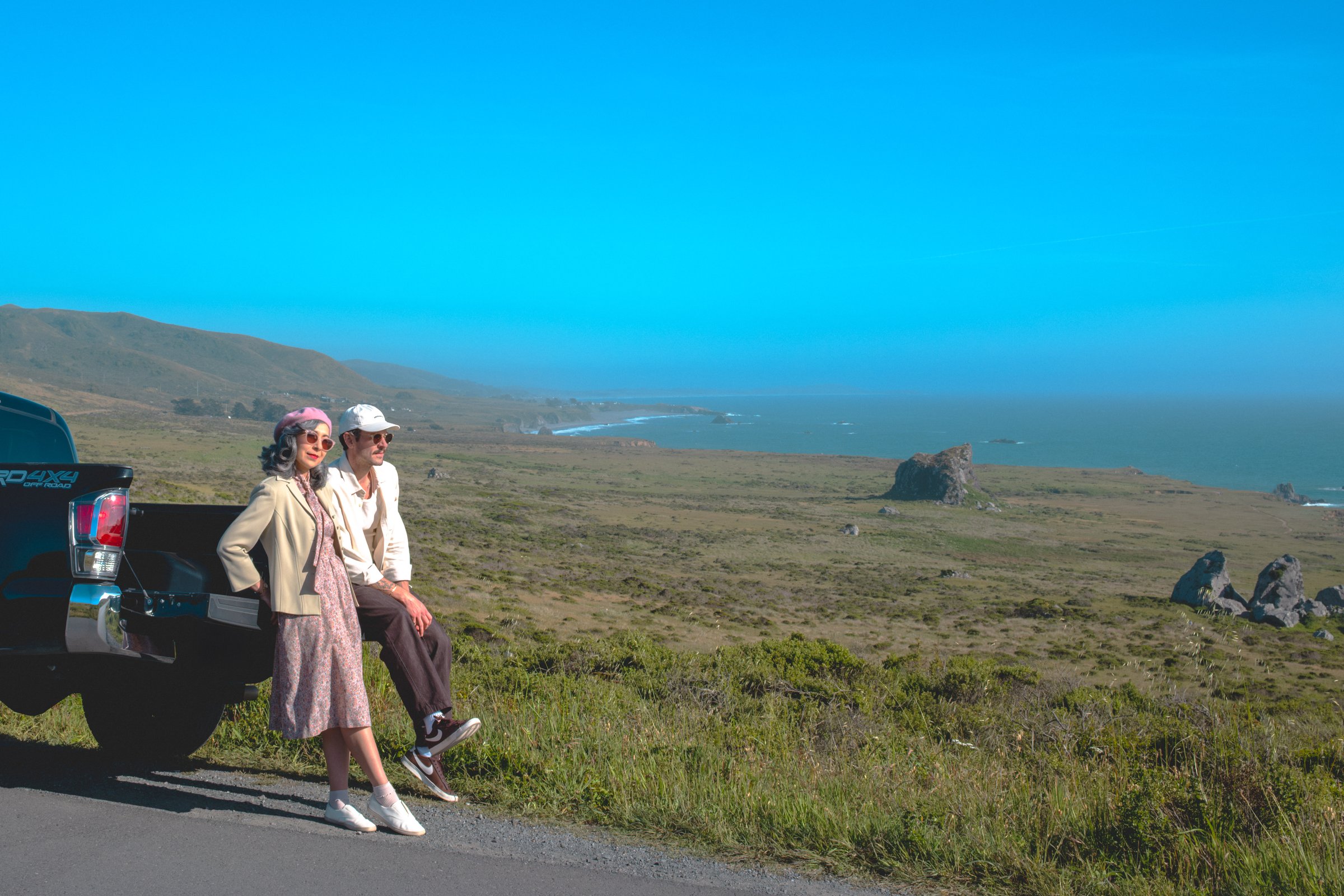 The Road to Bodega Bay gallery image: Two people are sitting on a black truck by a coastal road, overlooking a scenic view of a grassy landscape and the ocean under a clear blue sky.