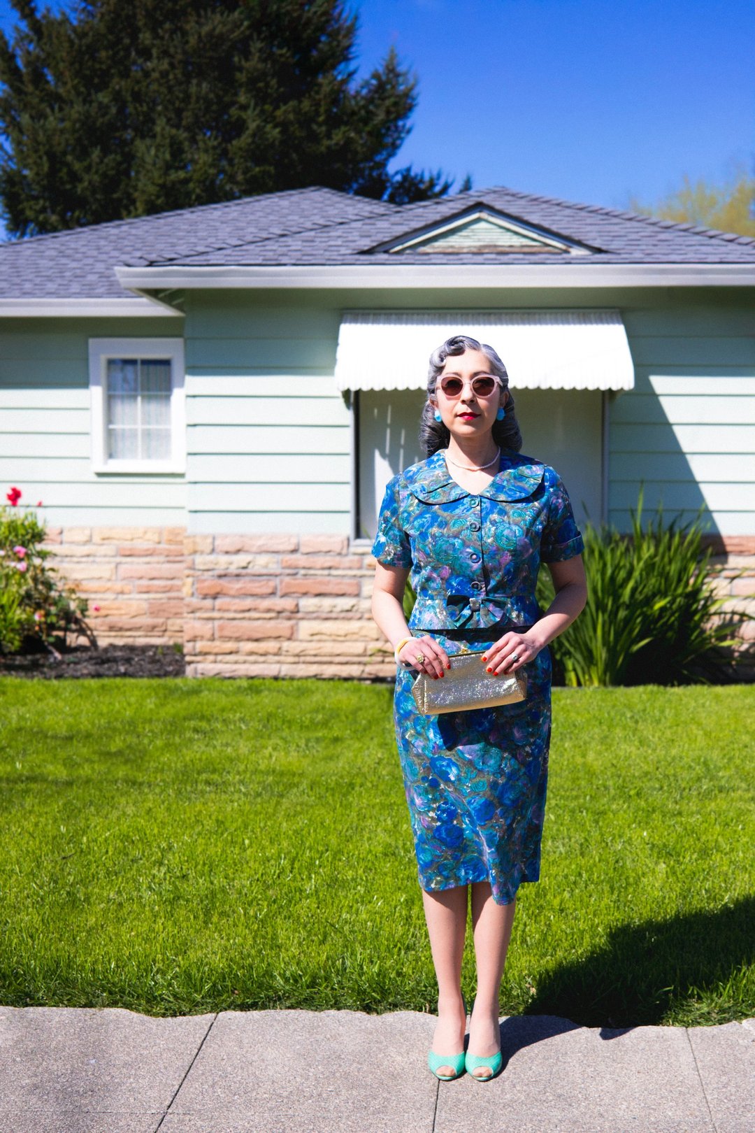 A woman in a floral dress and sunglasses stands in front of a light-colored house with a well-maintained lawn.