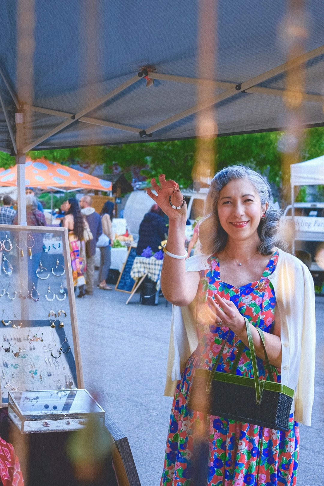 A woman in a colorful floral dress stands at a market stall, holding up a bracelet with a cheerful expression.