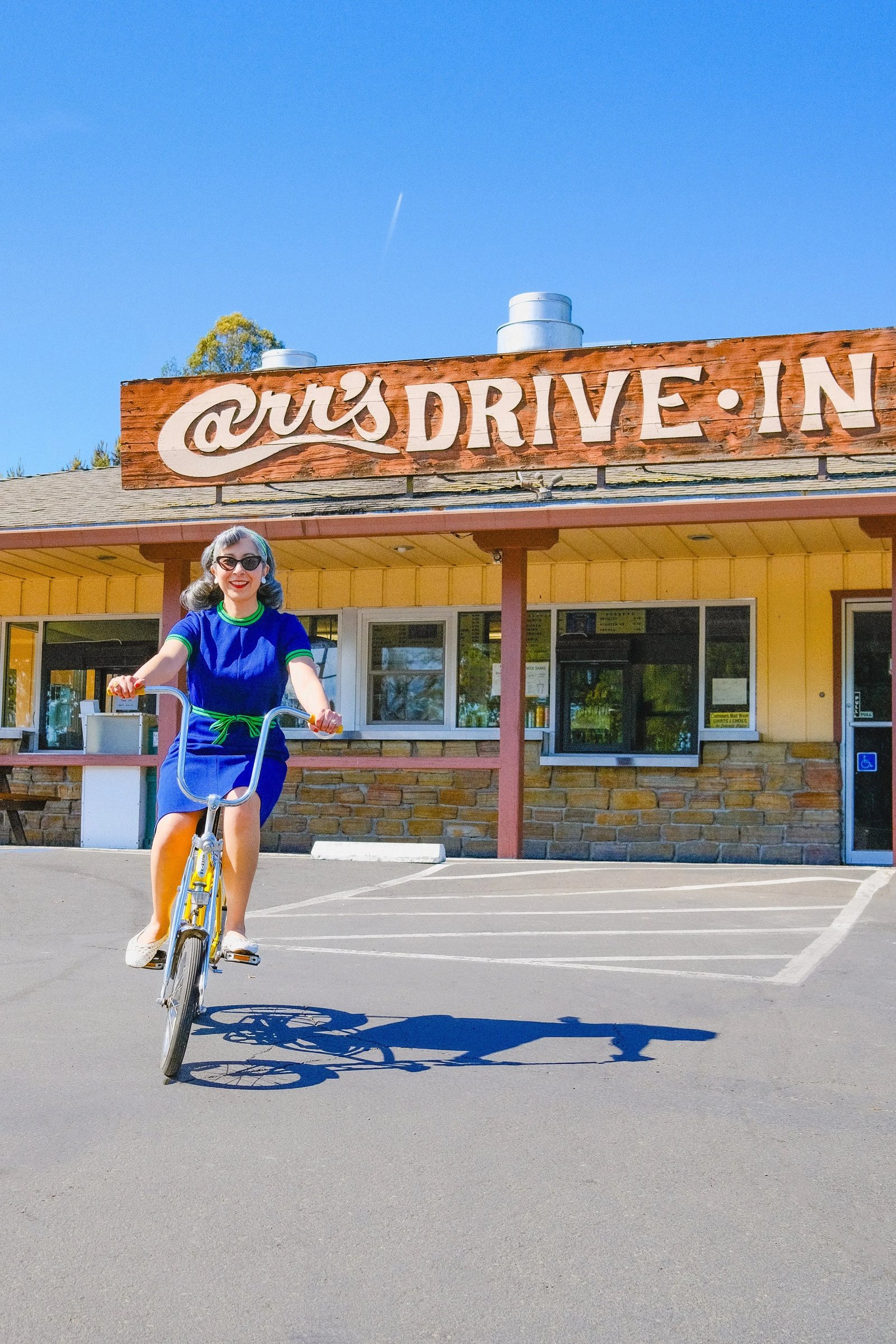 A woman rides a yellow bicycle in front of a retro drive-in restaurant sign.