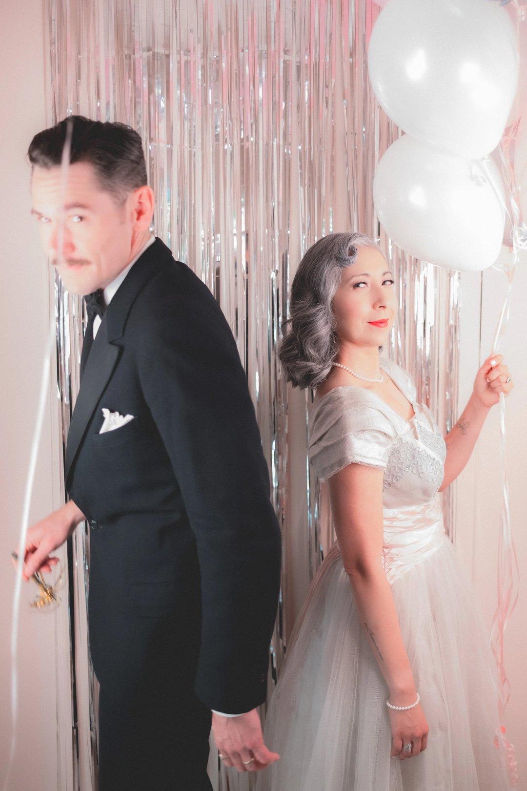 A man in a tuxedo and a woman in a vintage gown hold balloons and stand back-to-back in front of a shimmering backdrop.