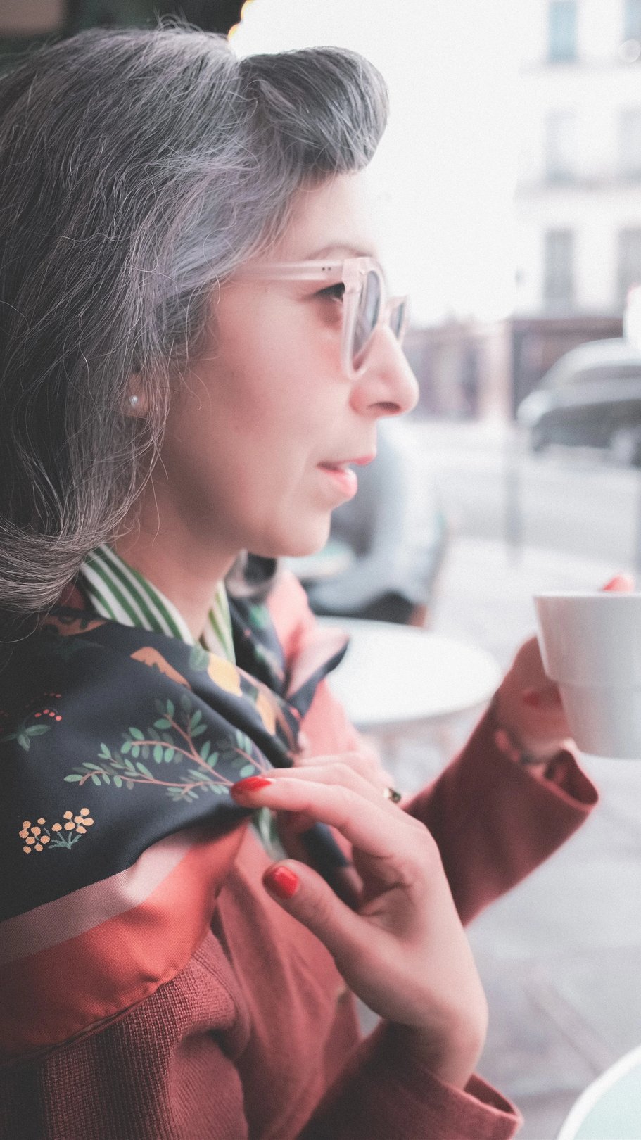 A woman with long gray hair and glasses holds a white cup while adjusting her patterned scarf at a cafe.