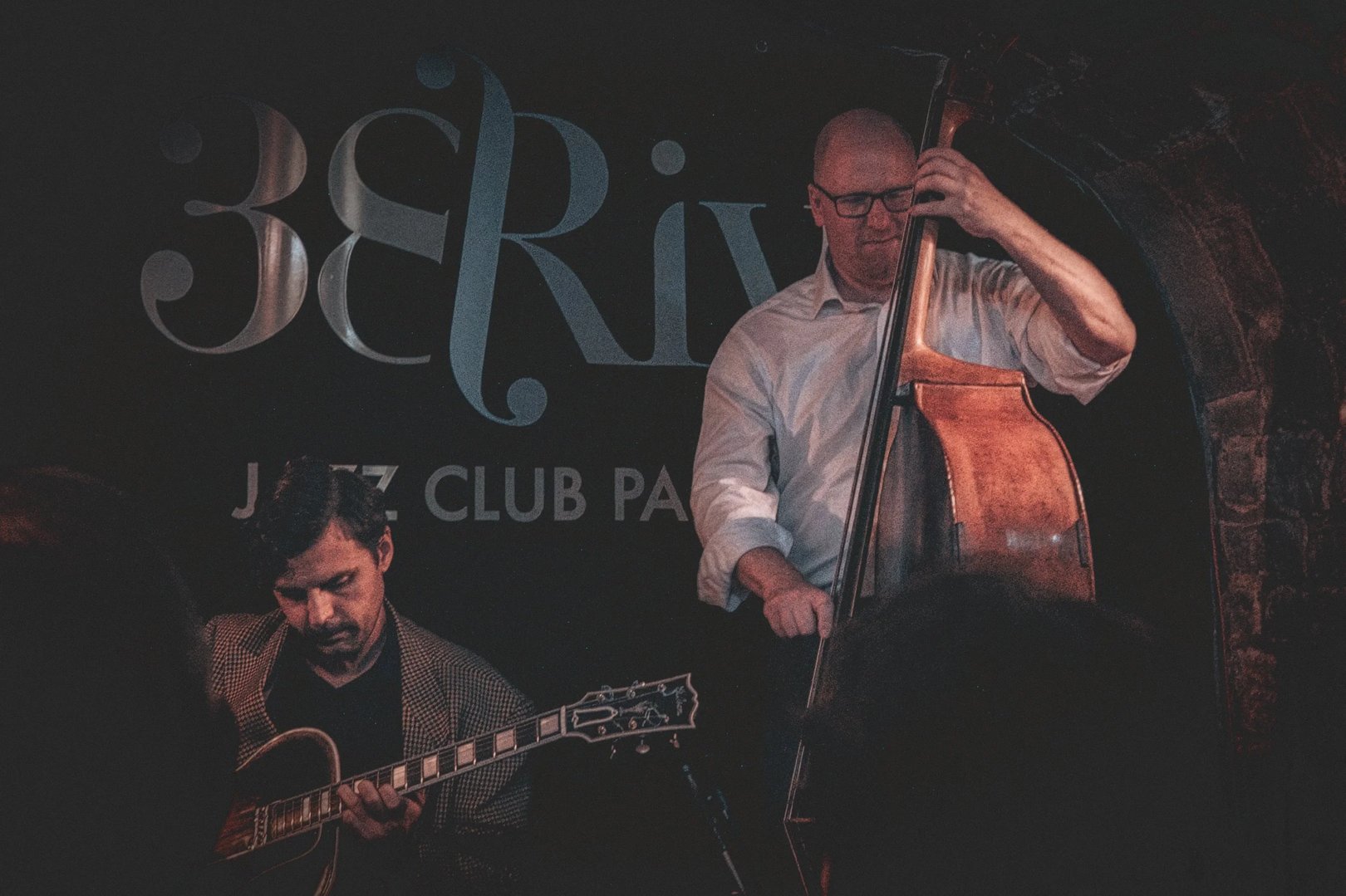 Two musicians perform on stage at a jazz club, with one playing the guitar and the other playing the double bass.
