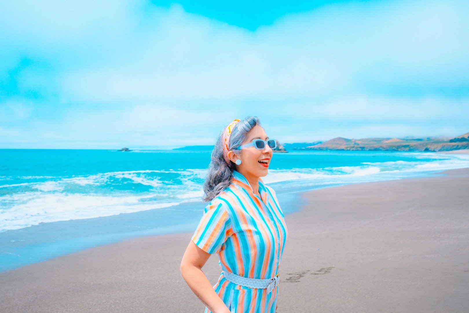 A woman in a striped dress and sunglasses smiles while walking along a beach with a bright blue ocean in the background.