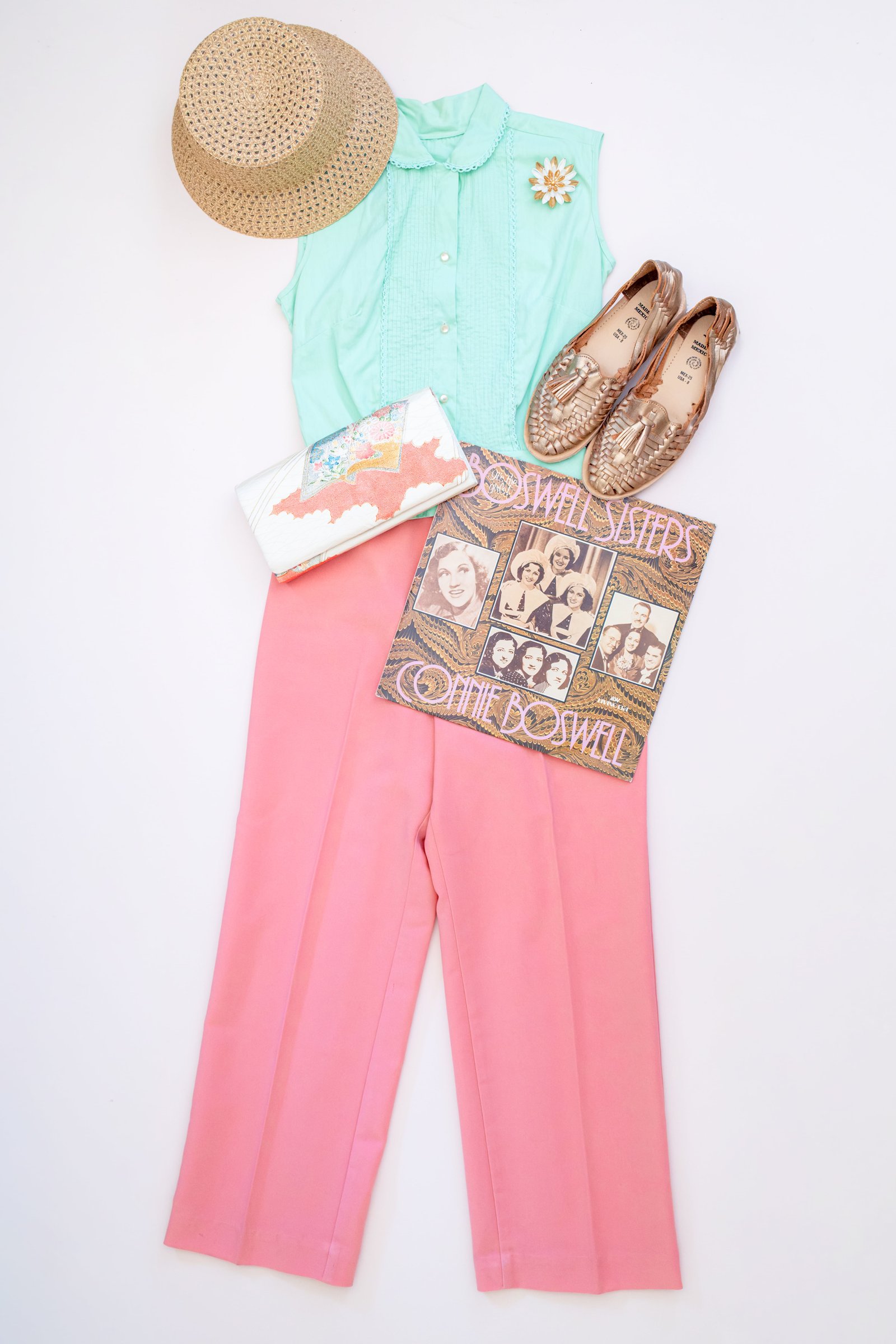 Inspired by Summer, Styled by Music gallery image: A summer-inspired outfit featuring a mint green blouse, pink wide-leg pants, a straw hat, bronze loafers, a floral clutch, and a vinyl record of The Boswell Sisters.