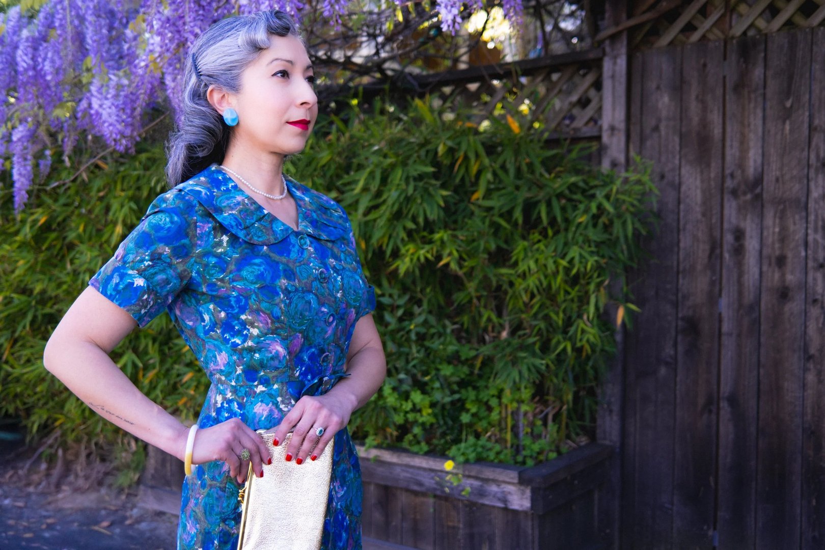 A woman in a blue patterned vintage dress stands elegantly outdoors, accessorized with jewelry and a clutch bag.