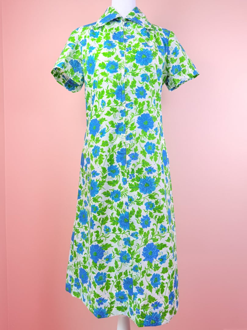 A 1960s women's blue flower print dress with a white background and short sleeves, displayed on a mannequin against a pink backdrop.