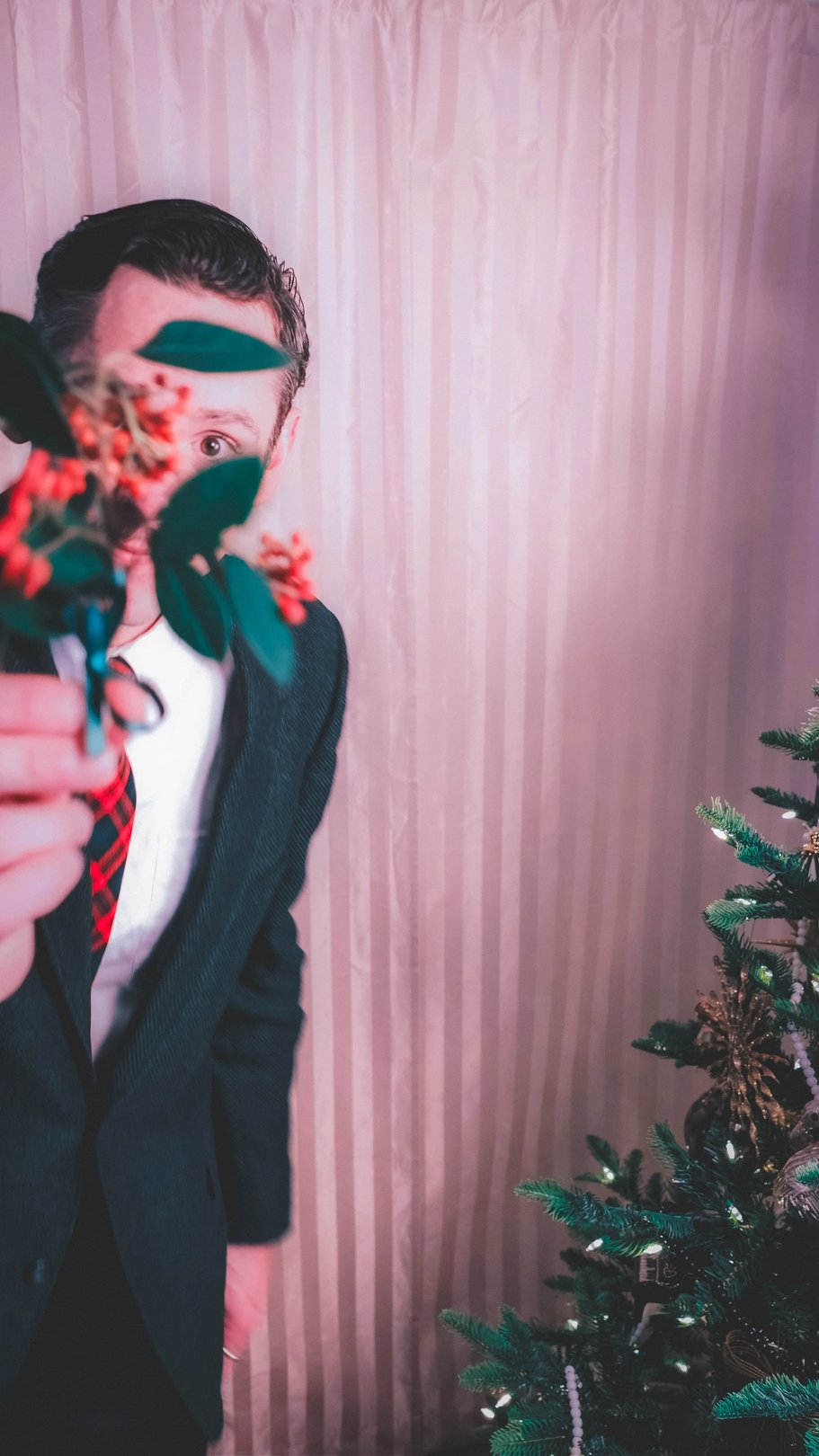 A person in formal attire holds a small bouquet of flowers in front of their face, with a decorated Christmas tree in the background.