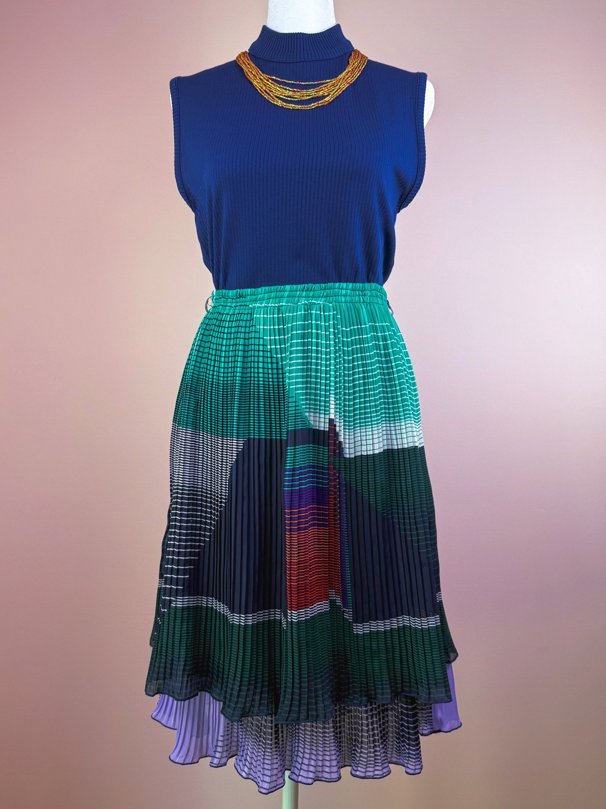 A women's 1970s green pleated skirt with a multicolored pattern, paired with a sleeveless blue top and a statement necklace.