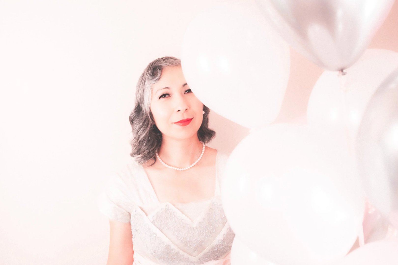 A woman with short, wavy hair and red lipstick stands against a light pink background, holding metallic and white balloons while wearing a vintage-inspired dress and pearl necklace