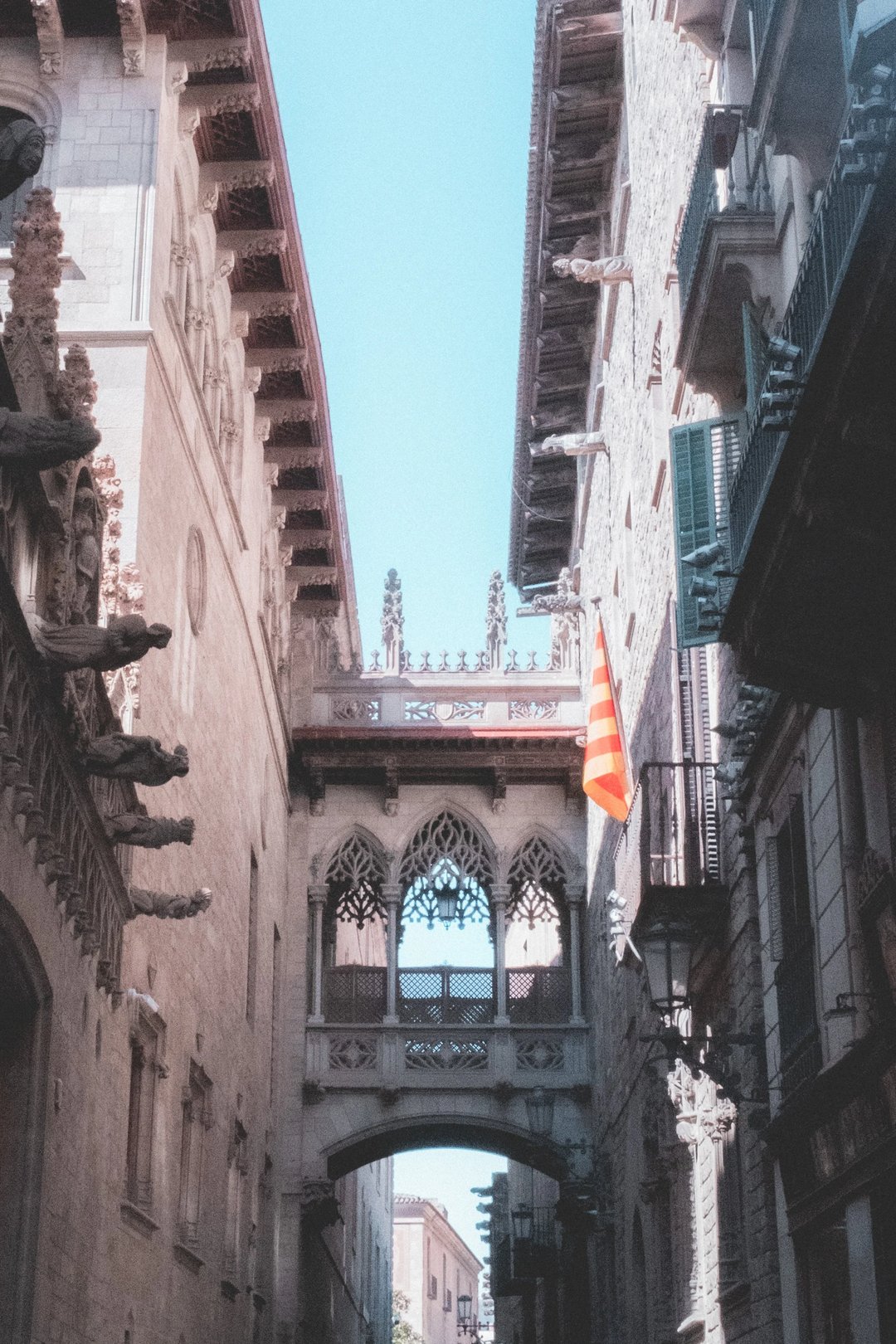 Narrow street in a historic city featuring a ornate arched bridge with intricate designs and a Catalan flag hanging.