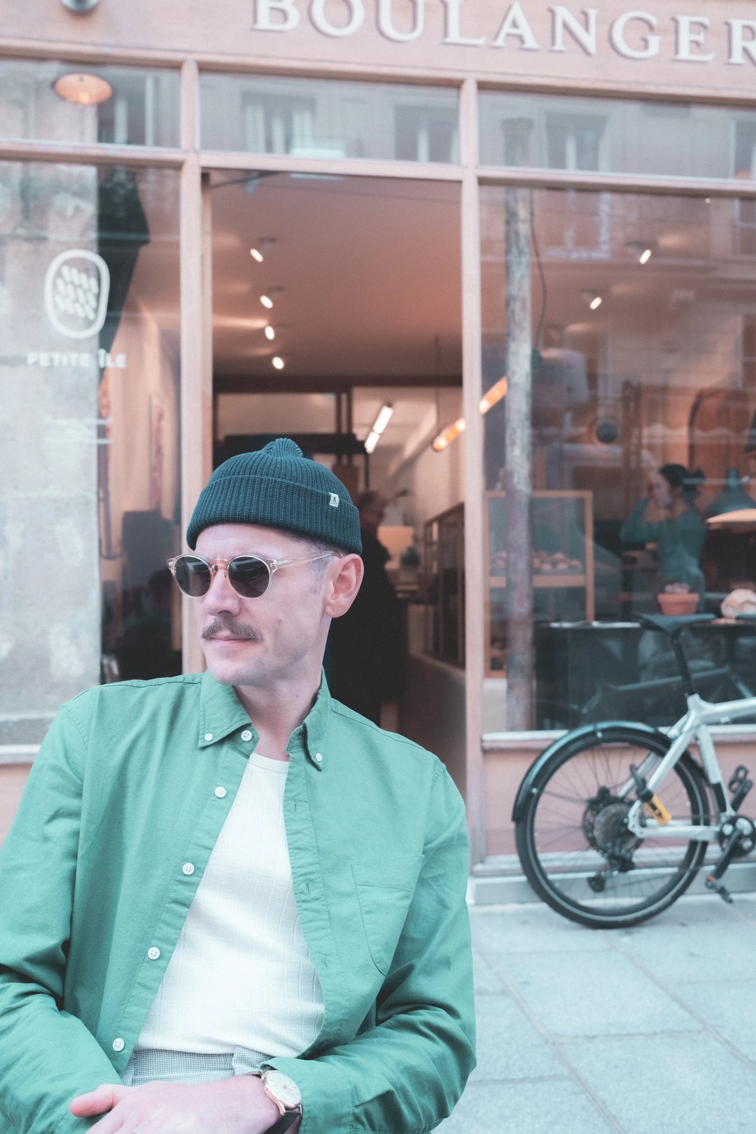 A man in a green shirt and black beanie sits outside a bakery, wearing sunglasses, with a bicycle nearby.