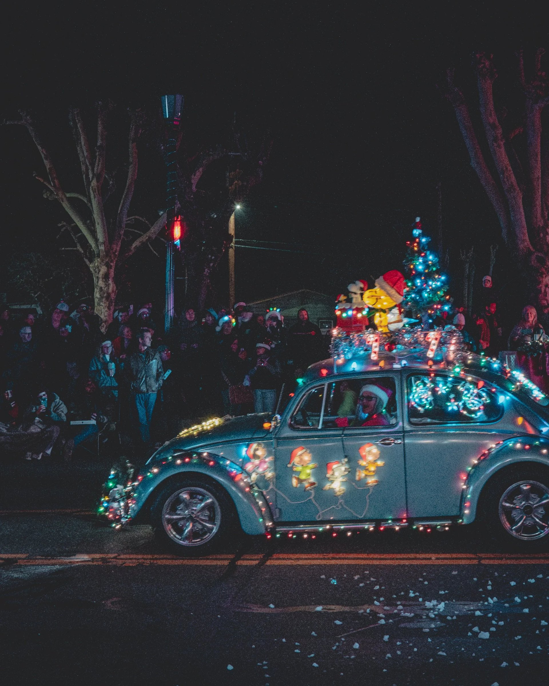 A decorated vintage car with Christmas lights and holiday figures drives past a crowd at a nighttime parade.
